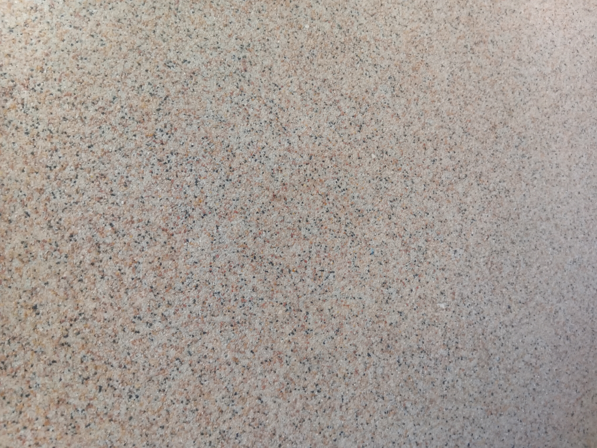 The image displays a close-up view of a textured surface, likely a wall or a ground. The surface is granular, composed of small, irregularly shaped particles in varying shades of beige, light brown, and black. The particles are densely packed, creating a uniform, speckled appearance across the entire frame. There are no visible people, objects, or distinct features that would indicate a specific location within the city of Verkhniachka, Ukraine, or any other environment. The lighting appears to be even, with no strong shadows or highlights, suggesting a neutral ambient light source.