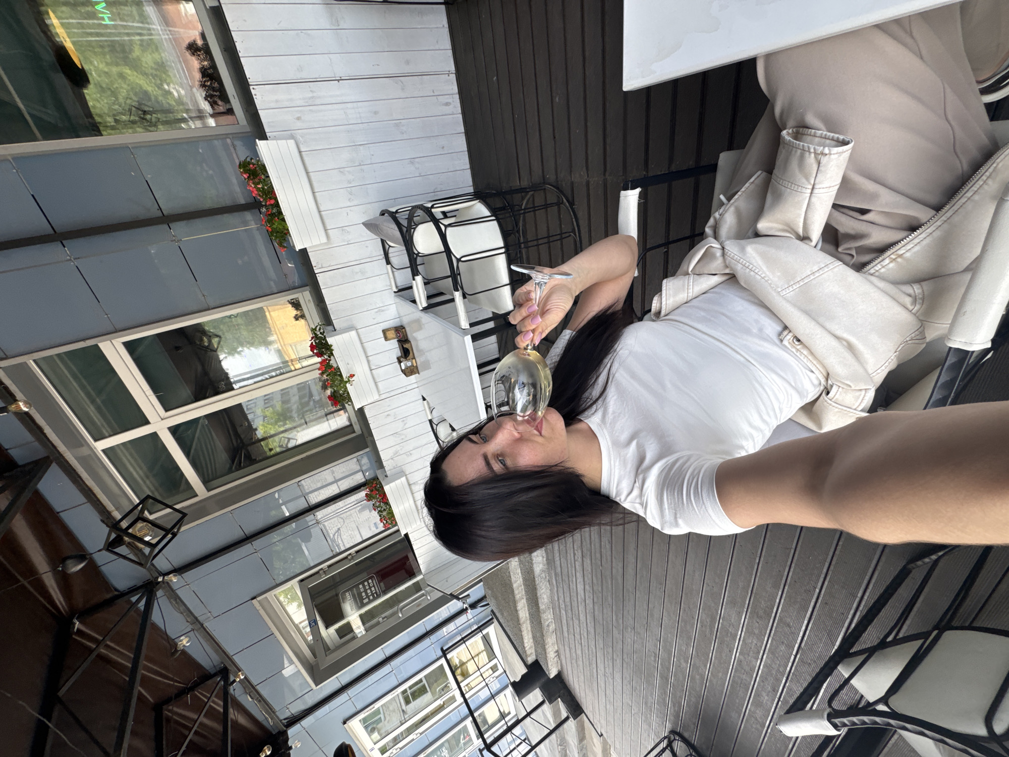 An eye-level shot captures a woman enjoying a drink at a semi-outdoor cafe or restaurant in Chaikovsky, Russia. The woman, with long dark hair, is wearing a white short-sleeved t-shirt and holds a wine glass containing a light-colored liquid, likely white wine, to her lips. Her nails are painted light pink. She exhibits a subtle smile, suggesting contentment as she poses for the photo.
The setting features dark wooden slatted flooring and contrasting white slatted wooden walls. Black metal chairs with white cushions are visible around white tables, some empty, and a light beige jacket is casually draped over a chair next to the woman. Flower boxes filled with vibrant red flowers adorn the white walls, adding a touch of color. Large windows enclose part of the space, reflecting greenery, other buildings, and the sky, indicating a bright daytime setting. Inside, decorative black metal light fixtures with exposed bulbs hang from above. The overall atmosphere appears relaxed and pleasant. Visible text in the reflections on the windows appears to be in Cyrillic, consistent with the location in Russia, though it is largely unreadable due to reflection and angle.
