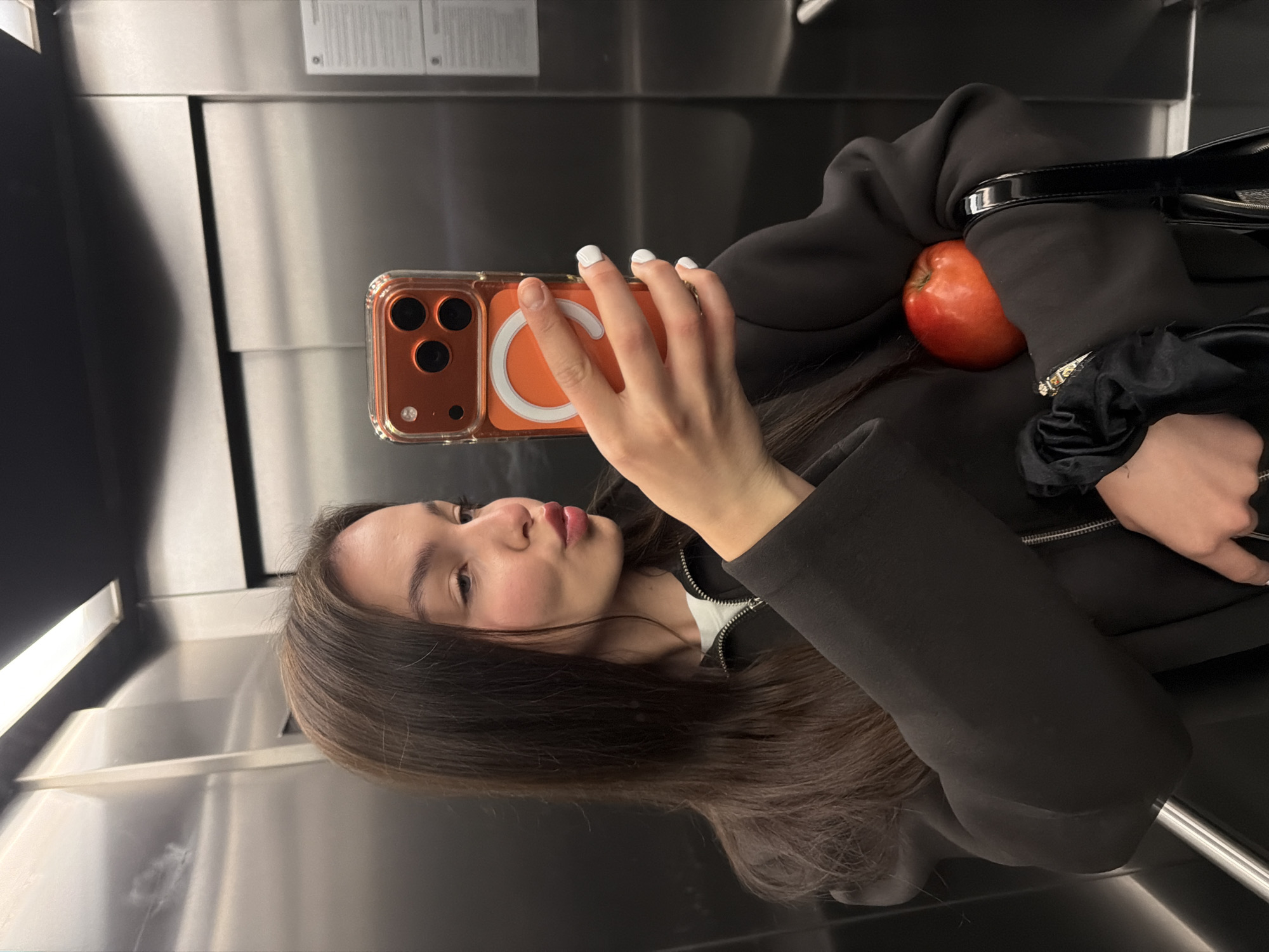 A young woman is taking a selfie in what appears to be an elevator. She is holding an orange smartphone with a clear case featuring a MagSafe charging ring. She has long brown hair, light skin, and wears white nail polish. She's dressed in a dark jacket. Next to her, an apple is visible on what appears to be a bag. The elevator walls are metallic and reflective. There is no clear indication that the location is in Sofia, Bulgaria.