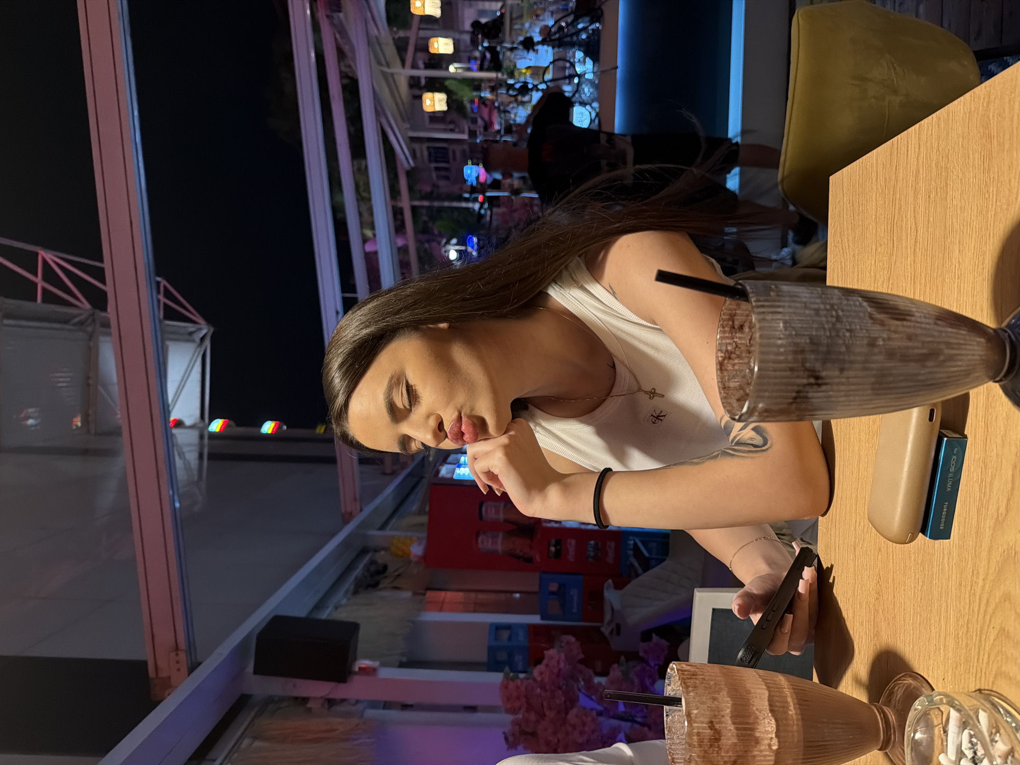 The image depicts a nighttime scene at an establishment in Plovdiv, Bulgaria, likely a bar or cafe terrace. A young woman with long dark hair is seated at a light wooden table, facing slightly to her right. She is wearing a white ribbed tank top with a "Calvin Klein" logo on the left chest and a crucifix pendant. Her eyes are closed, and her lips are pursed in a pronounced expression. Her right hand is positioned under her chin, and her left hand holds a black-cased smartphone. A black hair tie is visible on her right wrist, and a tattoo is present on her left upper arm.
On the table in the foreground, two tall, ribbed glasses contain dark beverages, possibly milkshakes, each with a black straw. Between the glasses, a clear glass ashtray holds multiple spent heated tobacco sticks or cigarette butts. Adjacent to the ashtray, a beige IQOS ILUMA device rests upon a blue box labeled "IQOS ILUMA TURQUOISE".
The background reveals an illuminated dining or bar area. To the woman's left, a portion of another person in a white shirt is visible, alongside pink floral decorations and stacked blue plastic crates. To her right, a bar counter with various bottles and a partially visible individual, likely a bartender wearing a black t-shirt and shorts, are illuminated by blue ambient lighting. The further background suggests an urban nighttime setting with blurred lights and structures. Overhead, a white and pink-hued retractable awning structure with metal beams and colorful decorative lights is visible.