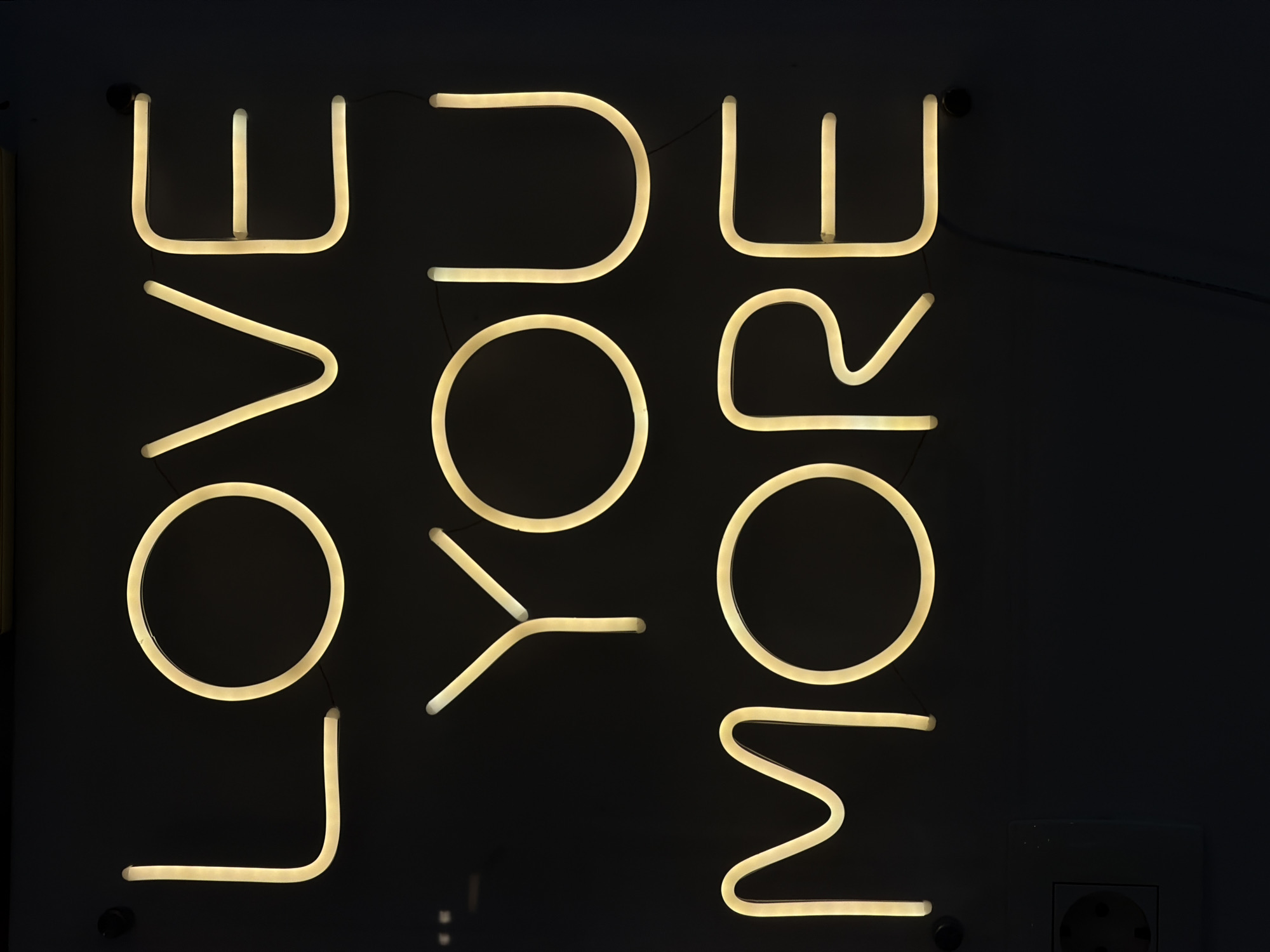 A glowing neon sign is prominently displayed against a dark background, likely a wall, creating a striking contrast. The sign spells out the phrase "LOVE YOU MORE" using three vertical lines of light-colored, possibly white or pale yellow, neon tubing. The word "LOVE" is on the left, "YOU" is in the center, and "MORE" is on the right, read from left to right and top to bottom. The letters are formed by continuous, smoothly bent tubes, emitting a soft, warm glow that illuminates the immediate surroundings slightly. The dark background emphasizes the brilliance of the neon lights. In the bottom right corner of the main image, a dark power outlet or switch is partially visible, indicating an indoor setting and the electrical nature of the sign. The overall scene conveys a feeling of affection and warmth, illuminated in a dimly lit or dark environment, suggesting either nighttime or an intentionally darkened space for display.