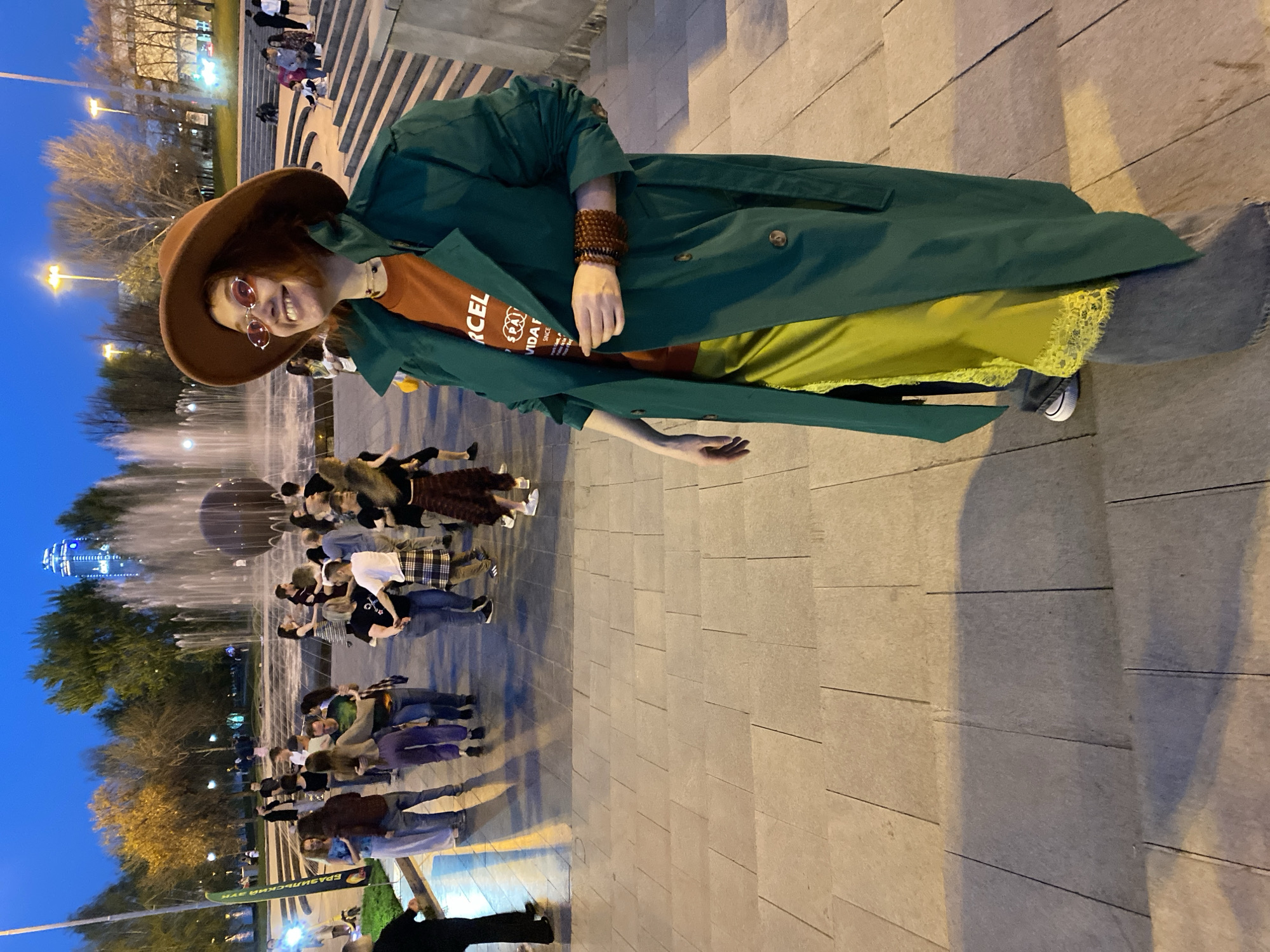 The image depicts a woman standing in a city square at dusk. She is wearing a brown hat, orange sunglasses, a rust-colored t-shirt, a green trench coat, a yellow dress with lace trim, grey pants, and grey sneakers. She also has beaded bracelets on her left wrist.
Behind her is a large fountain with numerous people gathered around it. Beyond the fountain, there are trees, street lights, and a tall building in the distance. The ground is paved with large rectangular stones.
The sign at the bottom left corner reads "ЕРАЗИЙСКИЙ ЗУБ", which transliterates to "EURASIAN TOOTH." This indicates the location is Yekaterinburg, Russia, where there is an architectural landmark known as the Eurasian Tooth.
