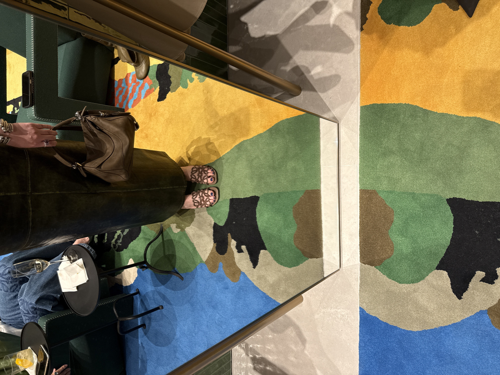 The image presents an overhead view of an indoor lounge or waiting area, likely in Zapopan, Mexico, characterized by a modern and upscale design. The scene incorporates reflections from mirrored surfaces, adding depth and complex visual elements.
**Subjects:** Parts of at least two people are visible. One person's legs and feet, adorned with brown patterned sandals and dark toe nail polish, are prominent, suggesting they are seated on dark green upholstered furniture. Another individual's hands and lower arms are seen holding a distinctive brown leather handbag, possibly a Loewe Puzzle bag, with silver hardware. This person wears multiple silver bracelets and a ring with a purple stone. A third person's legs, clad in blue jeans, are partially visible in the bottom left corner, also seated.
**Objects:** Key objects include a large, abstract rug featuring organic shapes in a vibrant color palette of yellow, various greens, blues, brown, and black. Dark green upholstered furniture serves as seating. Two small, black metal side tables are present; one holds an empty champagne flute and white napkins, while the other holds a glass with ice and a lemon slice, alongside a mobile phone. A long, shiny golden or brass handrail extends across the frame. The surrounding floor is made of light gray or beige tiles, with mirrored sections creating reflections. A black-cased smartphone is also visible resting on the arm of the green furniture.
**Activity:** The individuals appear to be relaxing in a social or leisure setting, possibly enjoying drinks. The presence of the handbag suggests they might be waiting, arriving, or departing.
**Notable Details:** The lighting is characteristic of an indoor environment, possibly evening or late afternoon, with artificial light casting subtle shadows. The overall atmosphere conveys a sense of relaxation and sophistication. No visible text is present in the image.