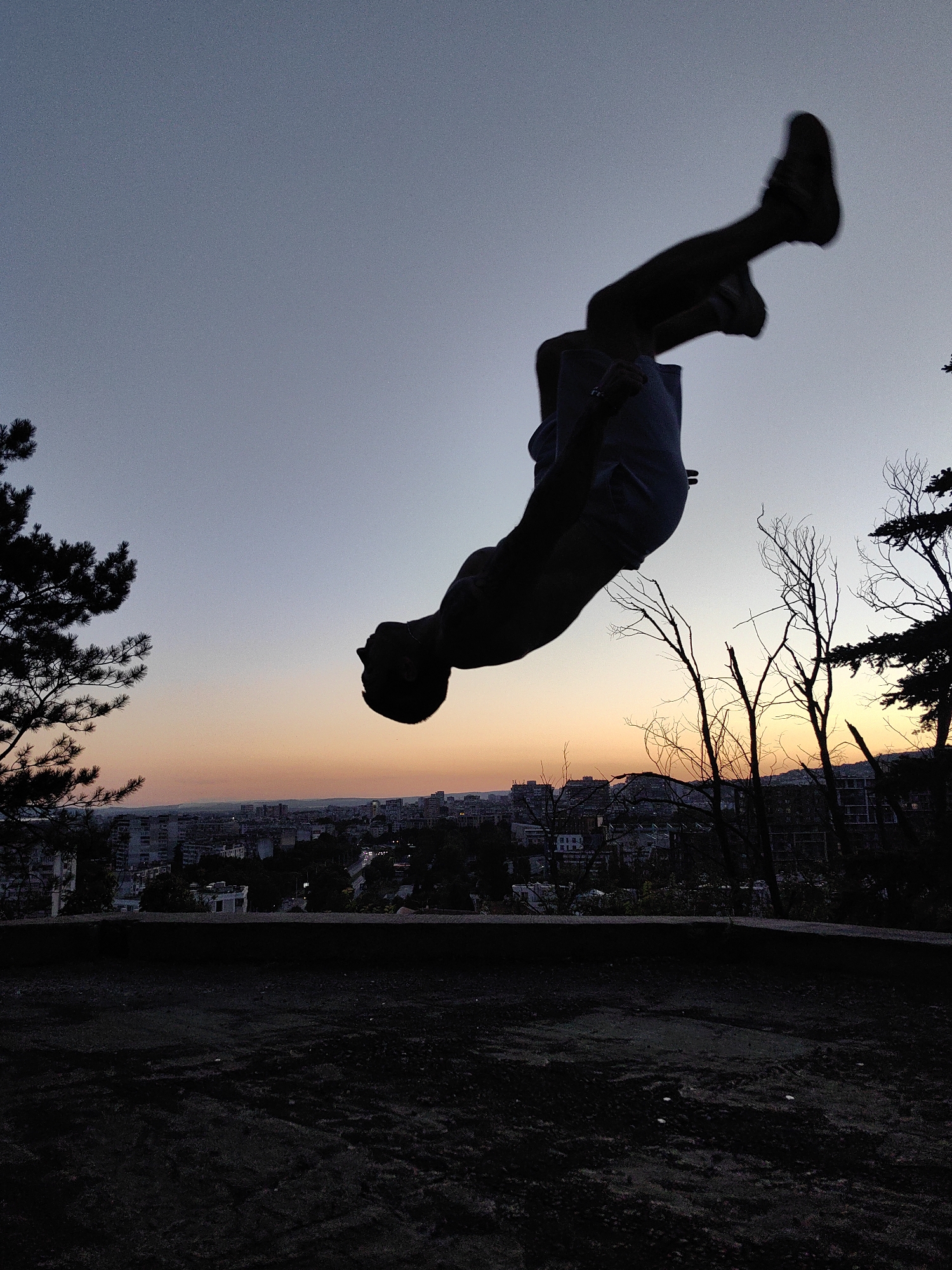 A silhouette of a person is captured mid-air, performing a backflip against a dusky sky with a gradient of blue to orange. The city of Varna, Bulgaria, is visible in the background, with buildings and lights appearing as dark shapes against the horizon. The foreground is dark and indistinct, suggesting a gritty, uneven surface. The image appears to have been taken at an elevated viewpoint, possibly overlooking the city from a park or a higher ground, like the "Asparuhov Most" (Asparuhov Bridge) or a park near the Sea Garden in Varna, given the panoramic view of the city and the silhouette of trees against the sky. The lighting suggests it is either dawn or dusk.