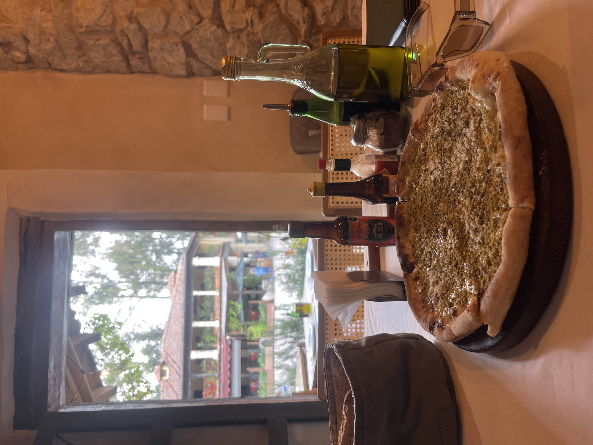 The image depicts a table setting inside a restaurant, likely in Domingo Arenas, Mexico, based on architectural styles visible through the window.
On the white tablecloth, there is a pizza on a dark-colored wooden serving plate, covered with green herbs or spices. Next to the pizza, there's a pair of sunglasses. There are also several bottles of olive oil and various hot sauces arranged in a row. To the lower left is a tan-colored breadbasket with bread inside.
Through the window, a view of buildings and trees is visible, including architectural details suggestive of a Mexican town. The scene is indoors, with stone walls visible behind the table setting.