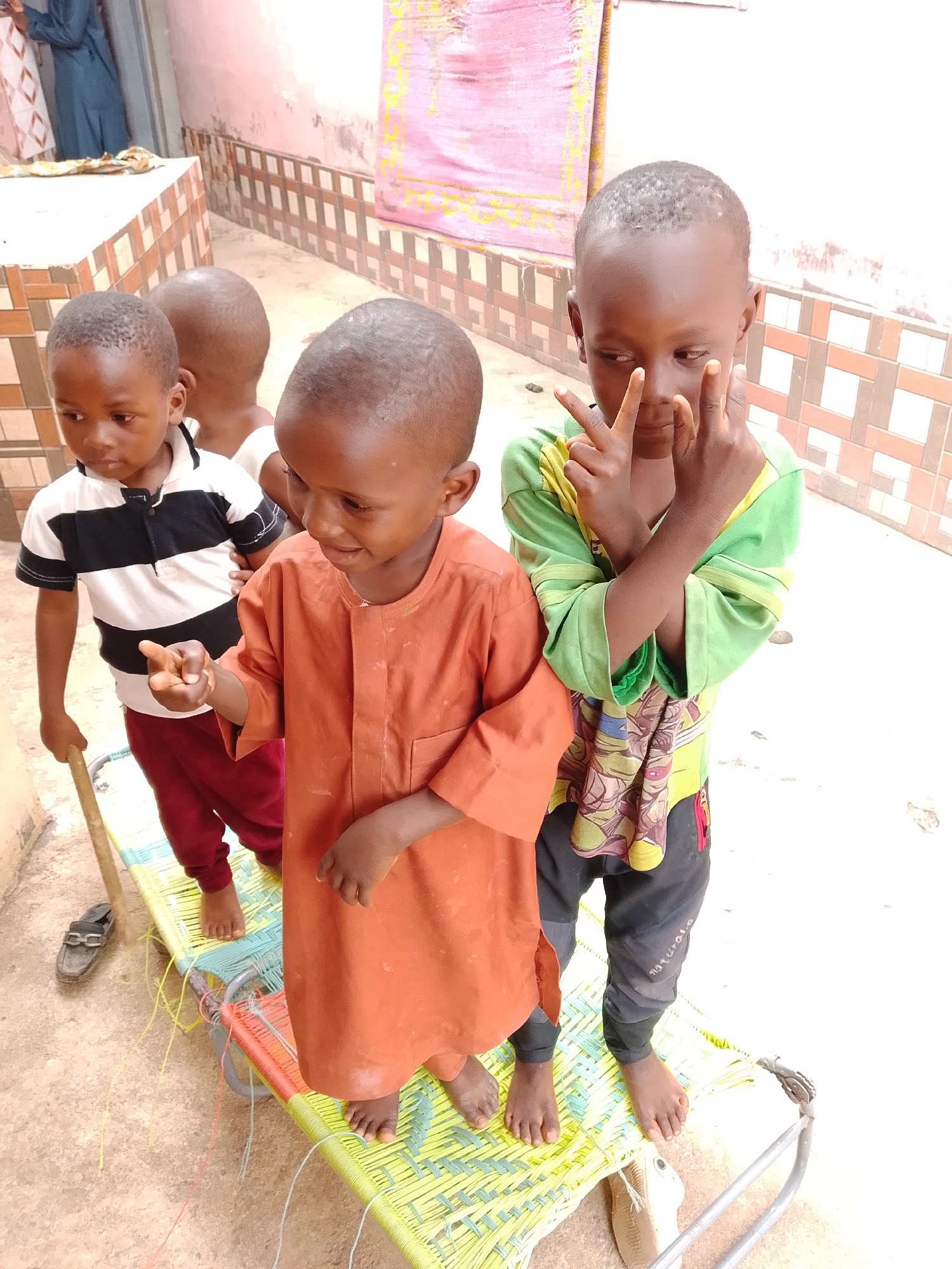 Four young boys are standing on a woven mat outdoors. The boy in the front right is wearing a green t-shirt and black pants, and is holding up two fingers on each hand in a peace sign gesture. The boy to his left is wearing an orange tunic and is looking down, pointing his index finger outward. Behind them, a boy in a black and white striped shirt and red pants is holding a wooden stick, and another boy is partially visible behind him. The background shows a tiled wall and a pink fabric banner. A black shoe is visible on the ground to the left of the mat.