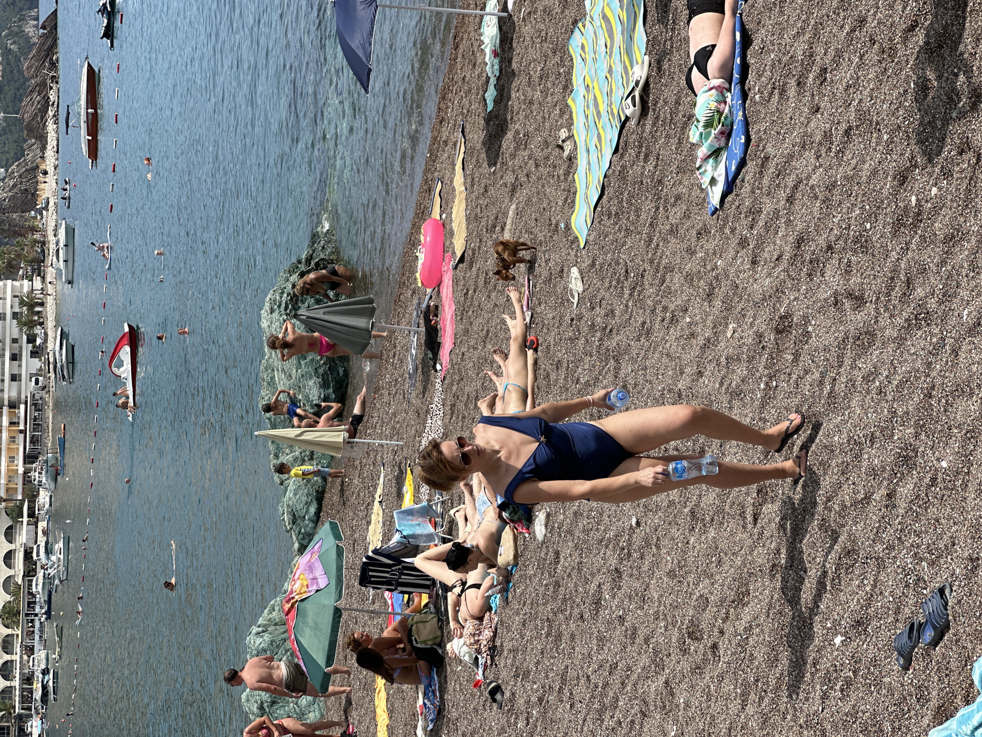 This is an eye-level, medium shot of a pebble beach in Rafailovići, Montenegro. In the foreground, a woman wearing a navy blue swimsuit walks towards the viewer, holding two plastic water bottles. She is wearing sunglasses and flip-flops. Behind her, several other people are sunbathing on towels, some lying down and some sitting up. A small brown dog is also visible on the beach, standing near a sunbather. In the background, the calm blue sea is dotted with various boats, including speedboats and smaller vessels. A waterfront promenade with buildings lines the distant shore, with rocky hills rising behind it. Beach umbrellas and towels are scattered across the beach, indicating a sunny day with leisure activities.