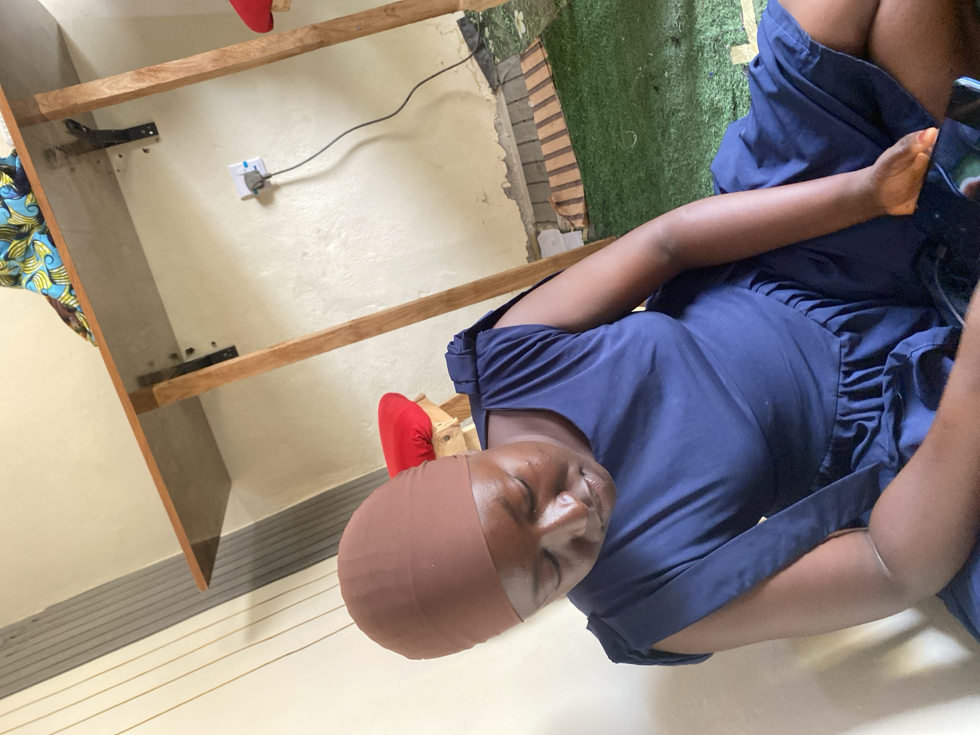The image depicts an indoor scene, possibly a residential setting in Jauro Yinu, Nigeria. A woman, wearing a dark blue dress and a brown headscarf, is seated on a green artificial grass floor. She is looking down, seemingly engaged with a mobile phone in her hands. The background reveals a light-colored wall with vertical grey and white striped paneling on the left. A wooden table, supported by wooden legs, is positioned in the upper right portion of the frame. A power outlet with a plug and cord is visible on the wall beneath the table. In the background, there are glimpses of red objects, possibly cushions, and colorful fabric on the table. The overall environment appears somewhat informal.