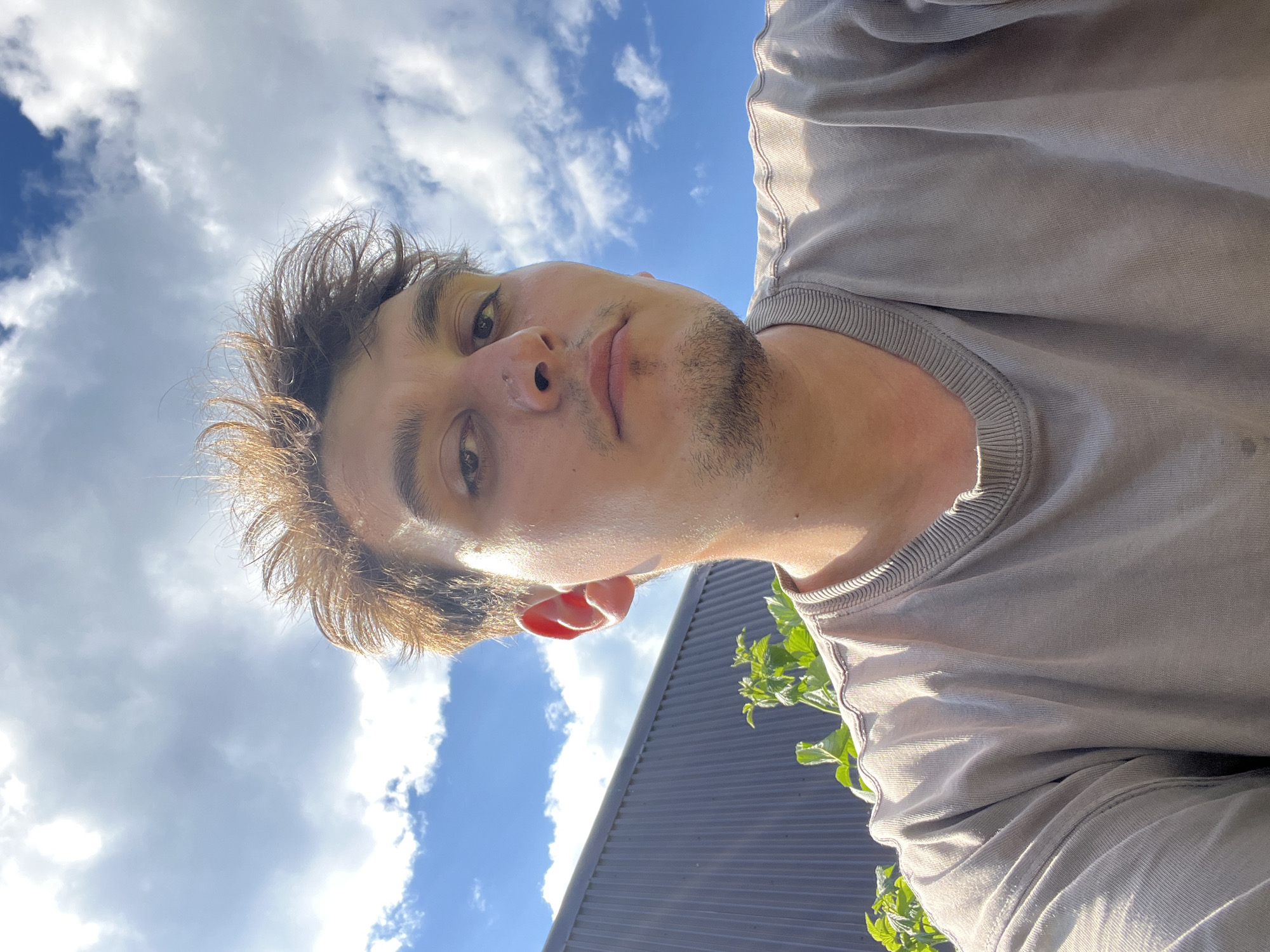 In a low-angle, outdoor, selfie-style shot, a young adult male with light brown, tousled hair and a light beard is depicted. He is wearing a neutral-toned t-shirt and looking directly at the camera. The background consists of a blue sky with scattered white clouds. To the left of the frame, a dark grey, corrugated metal structure is visible, with some green foliage in front of it. The lighting suggests it is daytime, with the sun casting a warm glow on the subject's face and the scene. The location is identified as Vasilyevo, Russia.