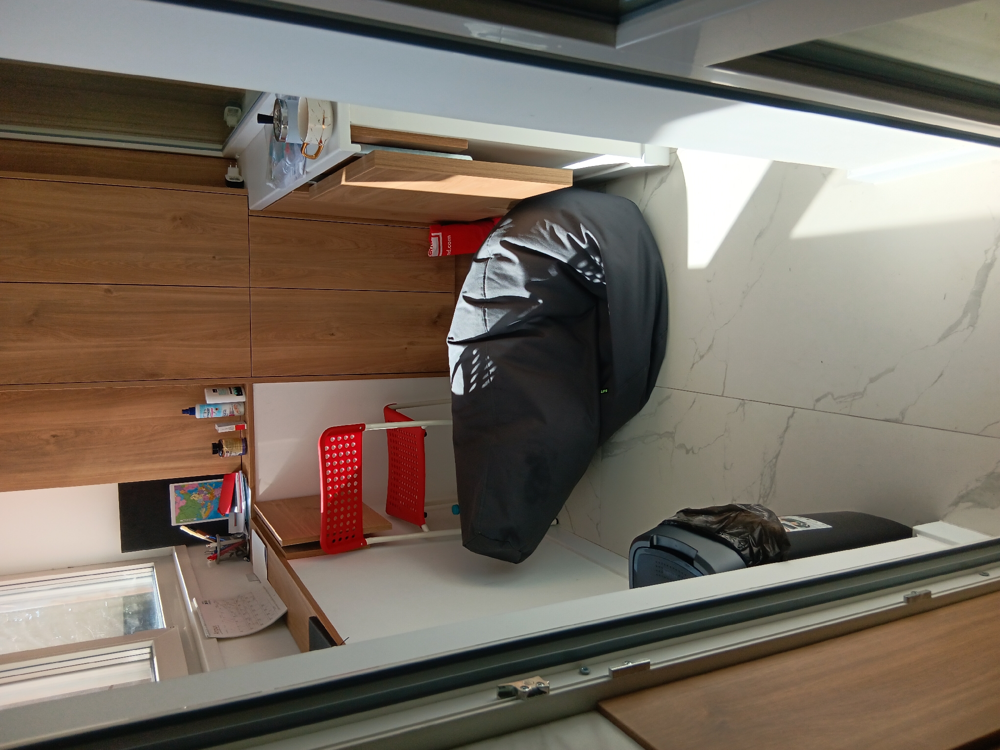 The image presents an indoor scene, likely a balcony or sunroom, attached to a room with wooden cabinetry. A gray, puffy jacket hangs partially over a red and white clothes drying rack positioned against a white, marble-patterned wall. A dark gray vacuum cleaner with a black plastic bag draped over it sits next to the wall. To the left, there's a desk or countertop area with stationary items, a window, and a small black chalkboard with a colorful map. The cabinets above the desk hold cleaning supplies and mugs. The frame of the balcony window is visible. The location is likely in Sofia, Bulgaria, but this is not definitively confirmed by the image alone.