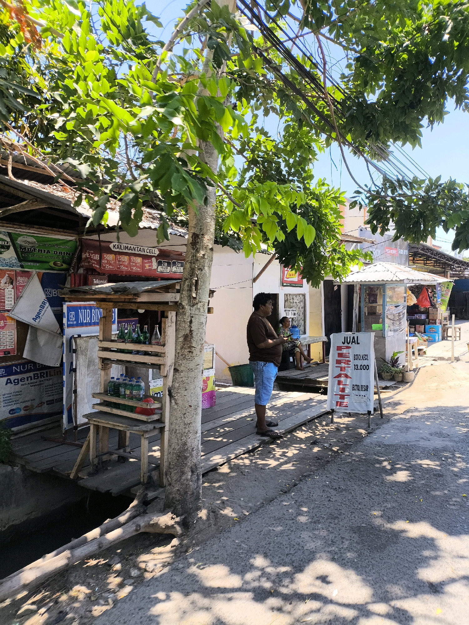 A man in a brown t-shirt and blue shorts stands on a wooden walkway next to a small wooden stand displaying bottles. The stand is set up in front of a building with several signs, including "MUTIHA DRINK" and "KALIMANTAN CELL." To the right of the stand, a white sign with red lettering reads "JUAL PERALTE BOTOL KECIL 13.000 BOTOL BESAR." The scene appears to be a street market or a small shop in a city. A second woman is visible sitting in the background. Overhead, tree branches with green leaves cast shadows on the ground. The sky is clear and blue. The overall environment is outdoor and appears to be during the day.