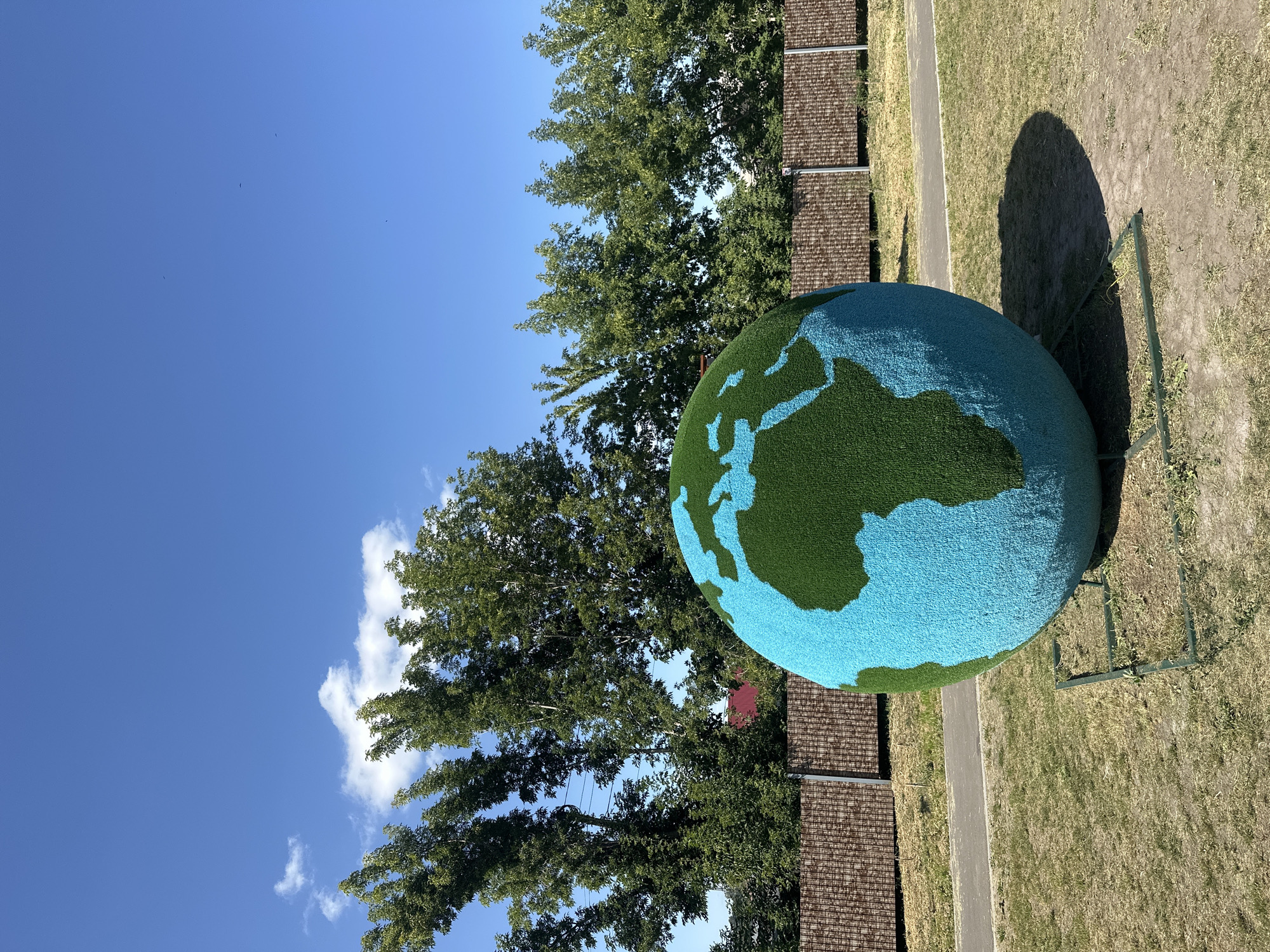 A large, artificial globe, representing the Earth with continents rendered in green and oceans in blue, is displayed outdoors on a grassy area. The globe is supported by a simple green metal frame. Behind the globe, a row of mature trees with dense green foliage forms a backdrop. Beyond the trees, a patterned fence made of vertical wooden slats in shades of brown and beige stretches across the scene. A paved pathway runs parallel to the fence, and beyond that, a small portion of a red-tiled roof is visible, suggesting buildings are present. The sky is a clear, vibrant blue with a few scattered white clouds. The sun casts a noticeable shadow of the globe onto the dry, patchy grass. The overall setting appears to be a park or public space.