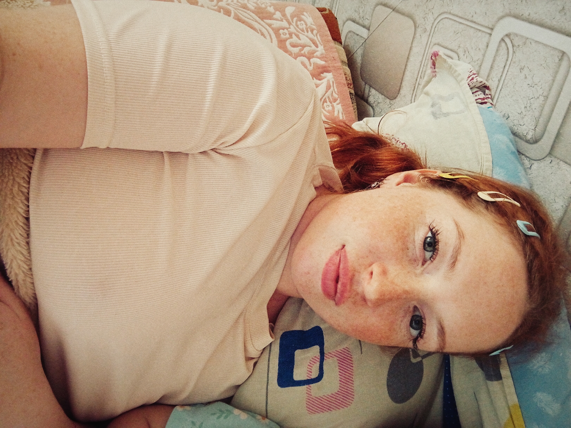 A red-haired, freckled woman lies in a bed in Unknown, Unknown. She wears a light-colored shirt and has several hair clips in her hair. Her head rests on a patterned pillow, and another pillow is visible behind her. The wall in the background has a textured pattern.