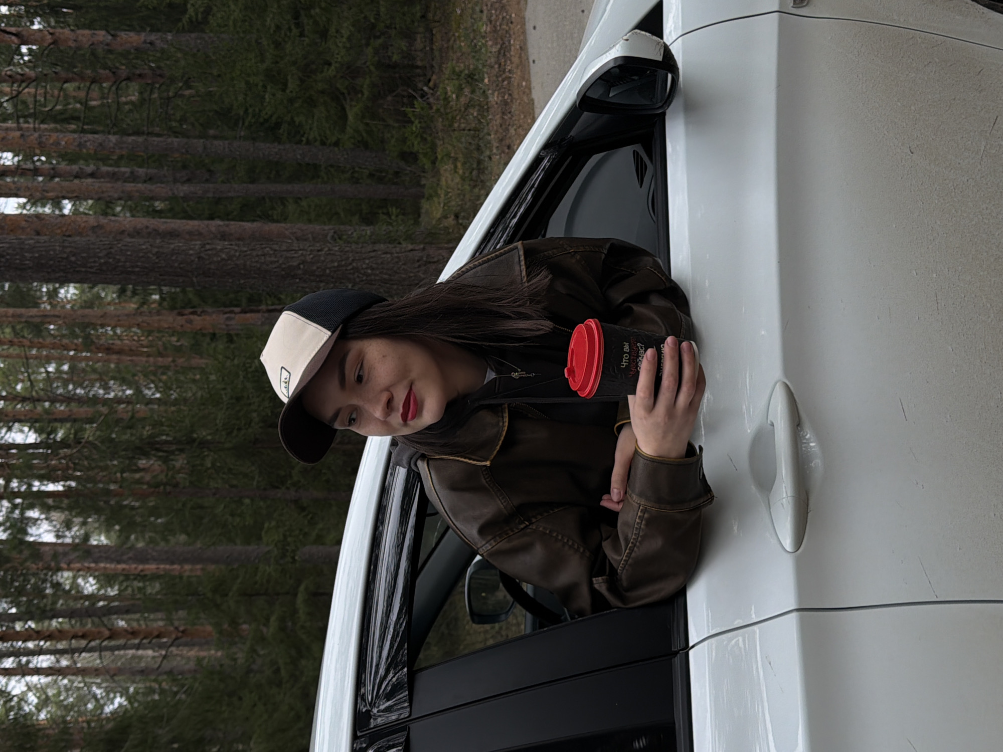The image captures a person leaning out of the passenger window of a white car, set against a backdrop of a dense conifer forest. The person, who appears to be a young woman, has long dark hair and is wearing a dark brown, possibly leather, jacket and a baseball cap with a black brim and a light beige front panel. She is holding a black travel mug with a bright red lid in both hands, looking towards the left of the frame with a soft, slightly smiling or thoughtful expression. Her lips are adorned with red lipstick.
The car is positioned next to a forest composed of numerous tall, slender trees, likely pines or firs, suggesting a natural and possibly remote setting. The lighting is diffused and overcast, indicating daytime without direct sunlight. The car's white exterior shows some minor marks or dust.
On the black travel mug, text in Cyrillic is visible, partially reading: "Что вы чувствуете сейчас?" (What do you feel now?). Below this, another word starting with "у" is partly visible, possibly "удивительно" (surprisingly).
The scene suggests a quiet moment of travel or contemplation amidst nature. The location context provided is Chaikovsky, Russia, which is consistent with the type of forest visible in the background.