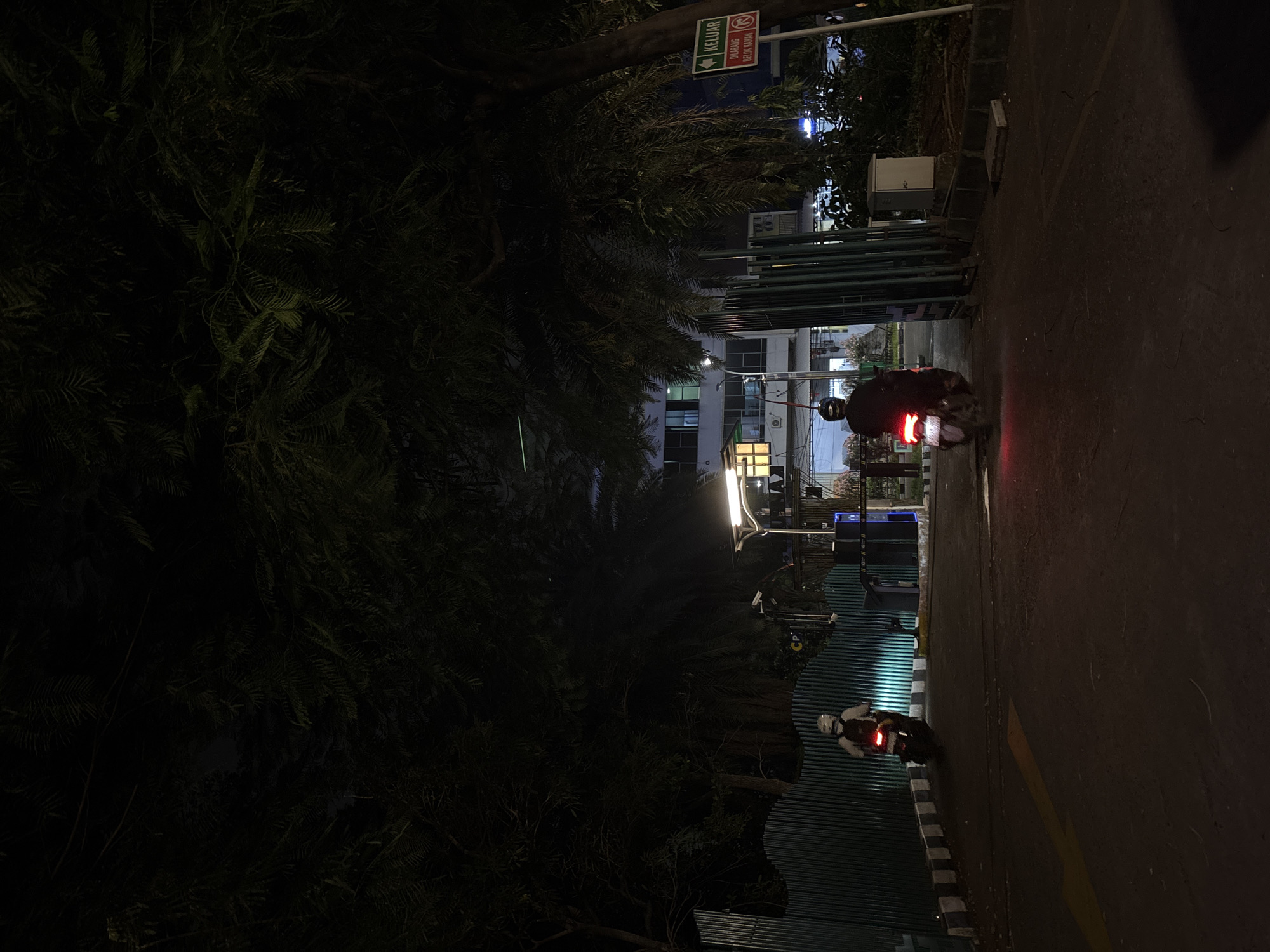 The image shows a street scene at night in North Jakarta, Indonesia. Two motorbikes are visible on the right side of the frame, with the taillights of one motorbike more prominent. A building is partially visible in the background, illuminated by artificial lights. Trees and dense foliage dominate the left side of the image. Traffic signs are visible near the top of the frame. The street surface is dark and reflective.