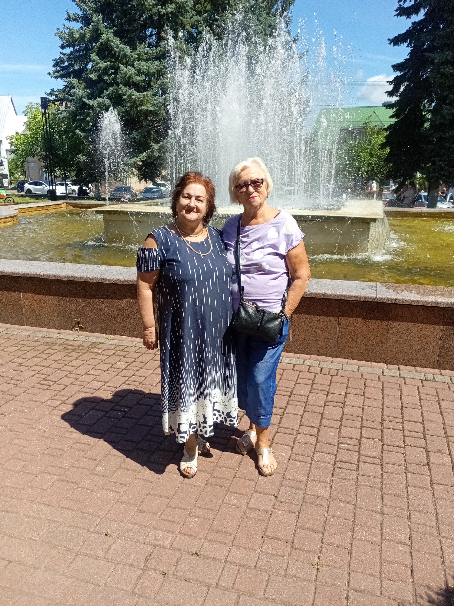 Two women stand in front of a fountain on a sunny day. The woman on the left is wearing a dark blue dress with white vertical stripes and white floral patterns on the hem. She has reddish-brown hair and is wearing a necklace. The woman on the right has blonde hair and is wearing glasses. She has on a light purple t-shirt with white heart shapes and blue jeans. She is carrying a black purse. They are standing on a brick-paved walkway, with the fountain behind them. The fountain has water spraying upwards. Trees and buildings are visible in the background. The location appears to be a public park or square in Tiachiv, Ukraine.
