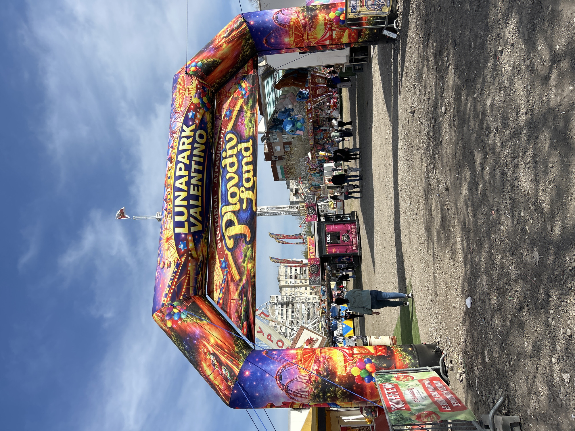 An outdoor scene captures the entrance to an amusement park or funfair in Plovdiv, Bulgaria, identified as "LUNAPARK VALENTINO" and "Plovdiv Land" by prominent text. The dominant feature is a large, colorful inflatable archway, adorned with images of fair rides, balloons, and stars, spanning a gravel path. The archway also displays the text "ФЕСТИВАЛ • АТРАКЦИИTE" (Festival • Attractions) and a date range "19-19 АПЕ" (likely April).
Beyond the archway, a bustling fairground is visible, featuring various attractions including a tall ride structure and a Ferris wheel-like ride in the mid-ground. Several fair stalls or booths are present, with one displaying "CASE" and "PINK DATE." The ground consists primarily of a textured gravel path, with a patch of green artificial turf near the arch.
Numerous people are seen walking through the fair, though most are partially obscured or in the distance. One individual in a dark jacket and jeans walks away from the camera in the foreground. The sky is bright blue with scattered white clouds, indicating clear weather and a sunny time of day, likely mid-day, with distinct shadows cast on the ground. The overall atmosphere appears festive and lively. Additional partially visible text includes "M", "YPO", "RRO", and "ТРАКИ".