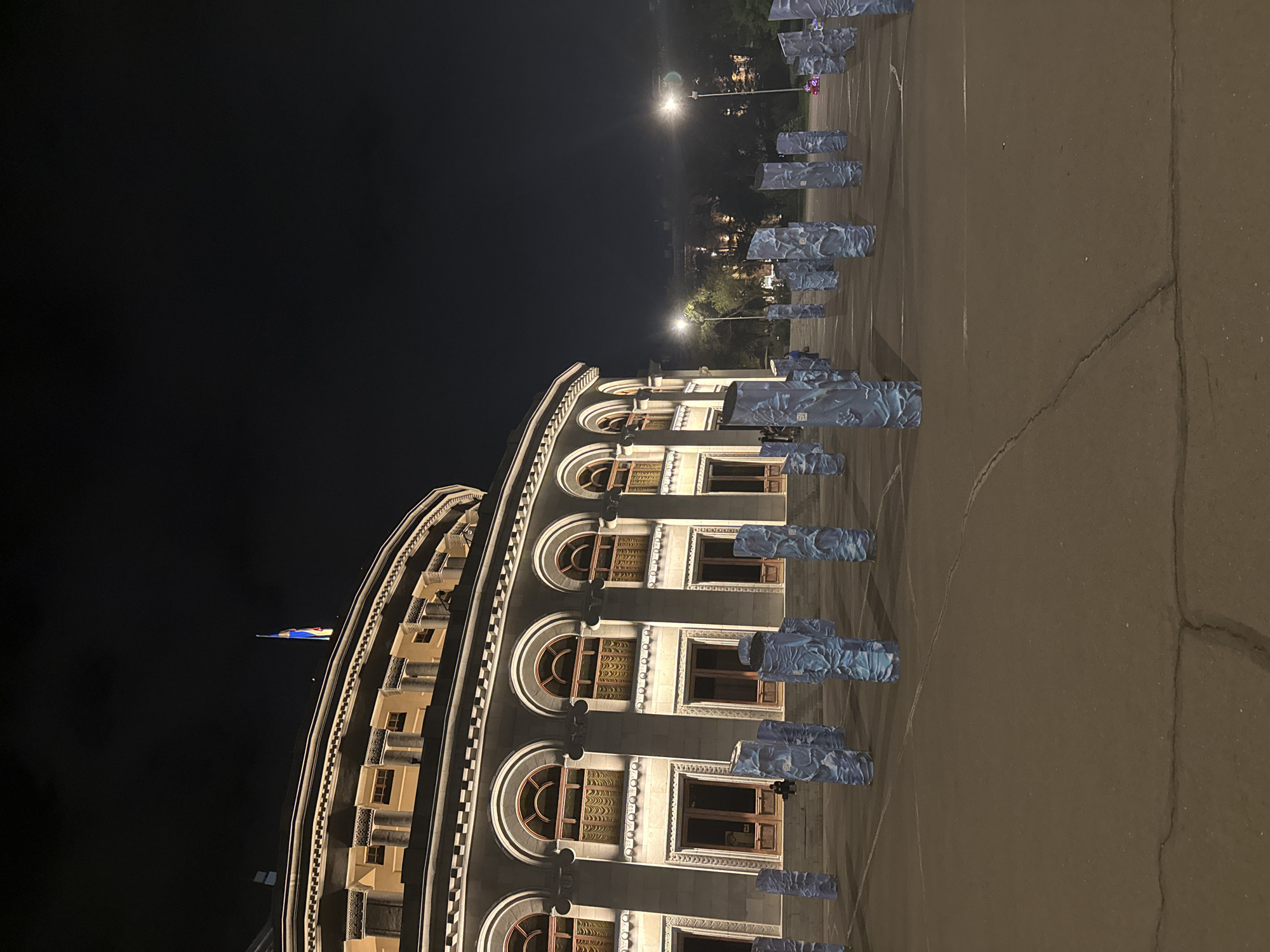 The image depicts a view of the Armenian Government Building located in Republic Square, Yerevan, Armenia, at night. The building is ornately decorated with a curving facade of arched windows and balconies. A flag flies atop the building. In the foreground, there is a paved area with multiple vertical rectangular panels bearing a pattern. The background includes street lights and other buildings in the distance.
