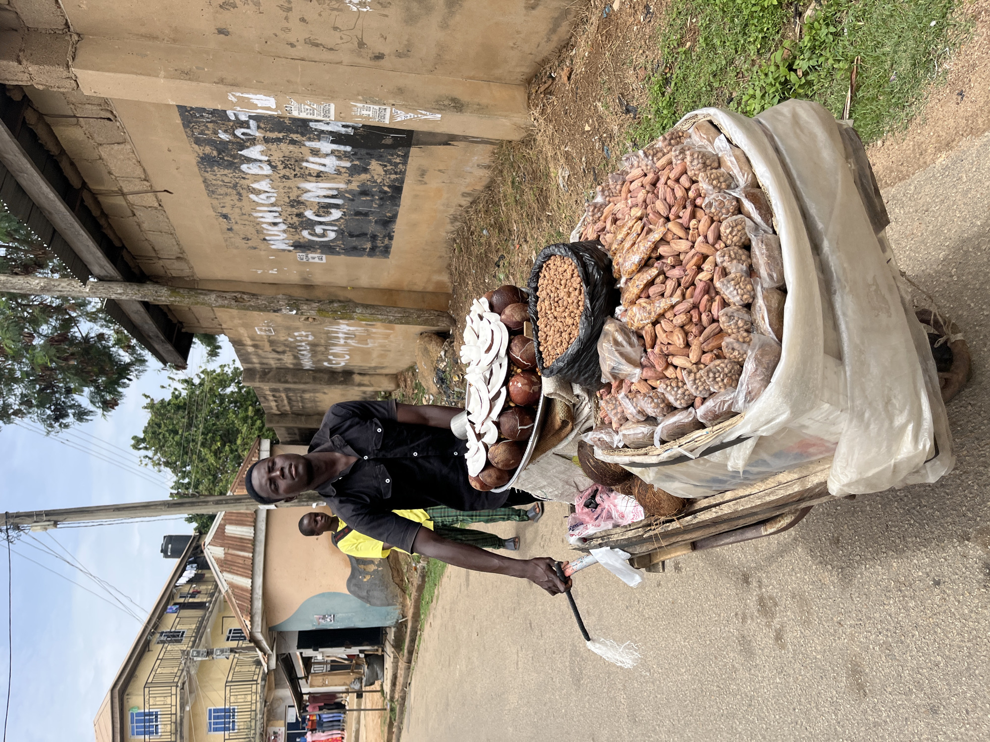 A man in Jos, Nigeria stands next to a cart laden with goods. The cart is covered in a white tarp and contains peanuts, groundnuts, and coconuts. The man is wearing a black shirt and holding a brush. The background includes a building with graffiti on the wall. Another man is walking behind the vendor, wearing a bright yellow shirt with black accents. The street is paved and there are buildings on either side. The sky is visible and partially cloudy.