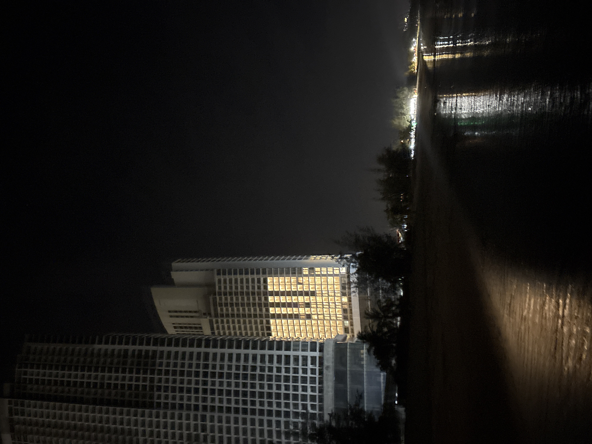 The image, when correctly oriented (rotated 90 degrees counter-clockwise), presents a nighttime view of a coastal or waterfront area, likely a city. On the left, two prominent modern high-rise buildings rise against the dark sky. The upper building features a distinctive facade with numerous windows, many of which are illuminated with warm yellow light, creating a bright, grid-like pattern across its upper stories. The lower building, partially visible beneath it, shares a similar grid-patterned architecture. Dark silhouettes of trees and vegetation are visible around the base of these structures.
A dark, undefined strip of land, possibly a beach or a promenade, runs horizontally across the middle ground, separating the buildings from the water. Some faint light reflects off this surface. To the right, a large body of water, likely the sea or a wide river, stretches into the distance. Its surface is dark but reflects numerous artificial lights from a distant shore or streetlights, appearing as shimmering, elongated streaks. The sky above is deep black, indicating late night.
The scene is tranquil and devoid of human activity. The primary illumination comes from the buildings' windows and distant lights reflecting on the water. The weather appears clear. There is no visible text in the image. The location context is Dong Hai Ward, Vietnam.