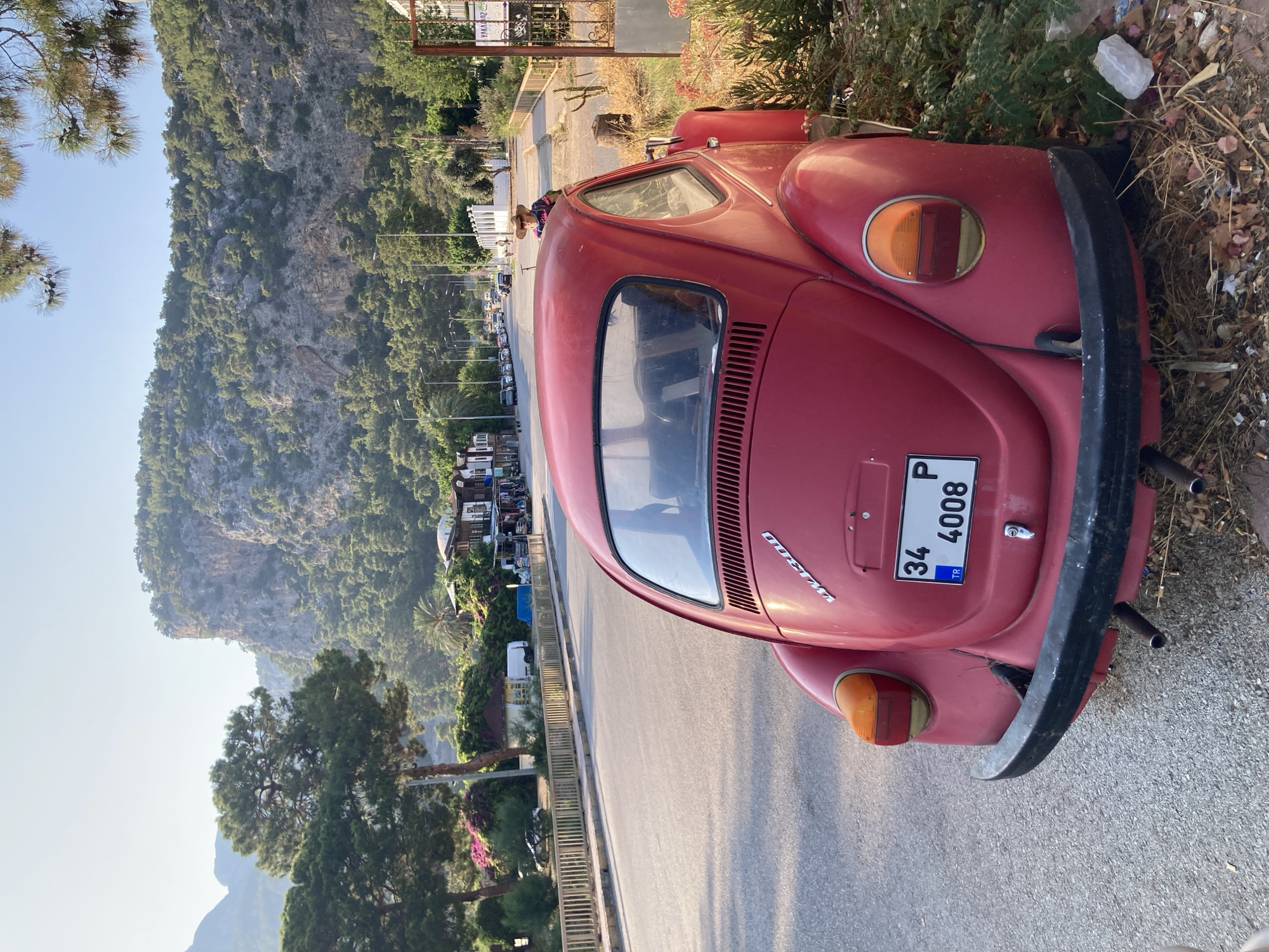 In Kemer, Turkey, a bright red Volkswagen Beetle is parked on the shoulder of a paved road, facing away from the viewer. The car's rear is prominently featured, displaying its taillights, chrome bumper, and license plate which reads "34 P 4008 TR." The car is situated on the right side of the road, with dry vegetation and some litter surrounding its tires.
The road runs parallel to a sidewalk lined with a light-colored railing and then a row of buildings, some appearing to be shops or restaurants. Behind these structures, lush green trees and palm trees are visible, leading up to a dramatic, rocky mountainside covered in dense forestation. The sky above is clear and a light blue, suggesting it is daytime.
In the distance, a line of cars is traveling along the road, and on the sidewalk, a person wearing a straw hat and a colorful top is visible, seemingly observing the scene. The overall impression is a roadside view in a scenic, possibly tourist-oriented area of Kemer, characterized by its natural beauty and the presence of classic vehicles.