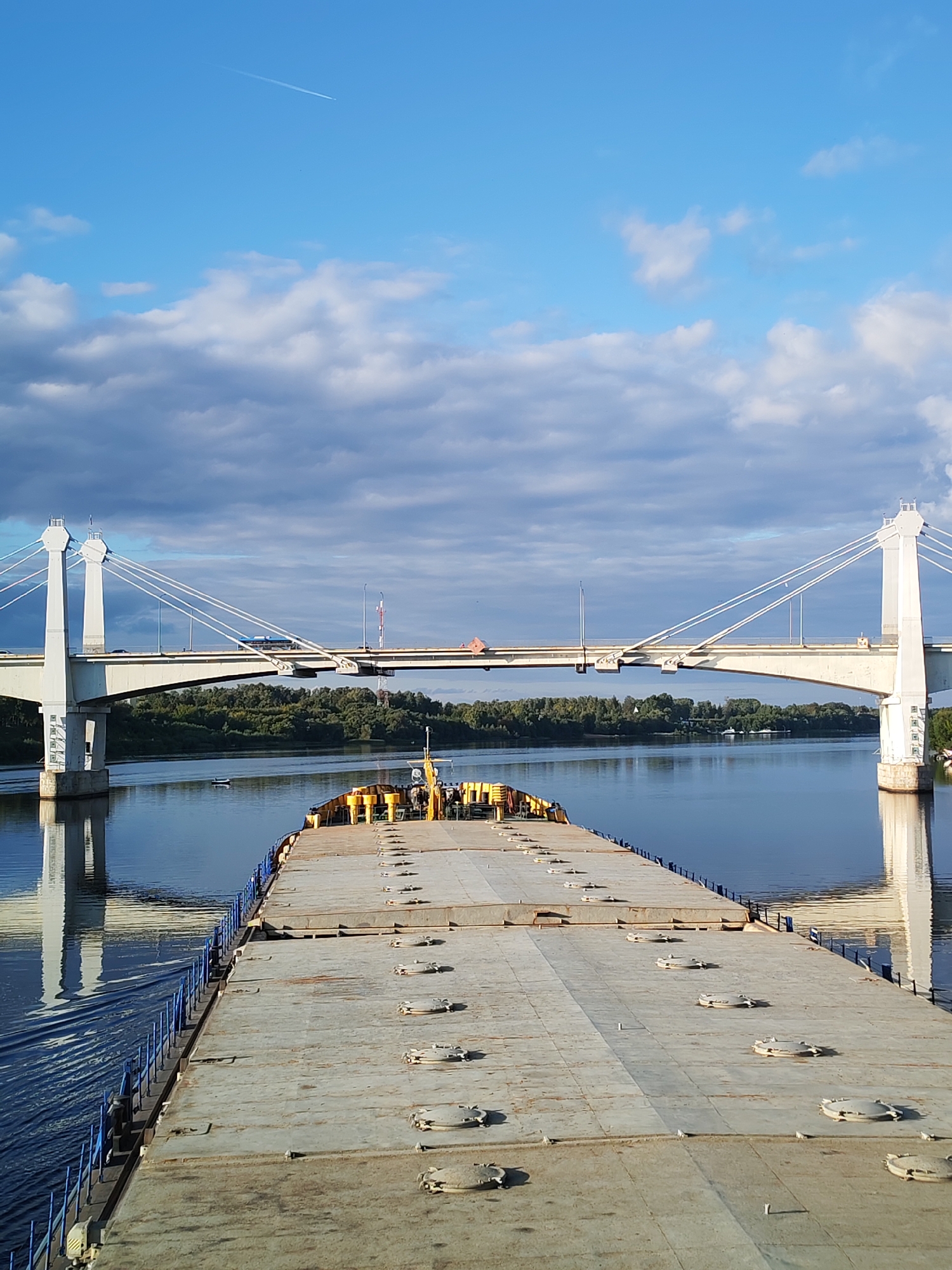 This image captures a scene from the Volga River in the city of Kimry, Russia, with a focus on a barge in the foreground and a cable-stayed bridge in the background. The barge, a large, flat-topped vessel, is positioned centrally in the frame, its deck covered in numerous circular hatches. The river itself is calm and reflects the sky and the bridge.
Dominating the background is a substantial cable-stayed bridge, characterized by its two large, white, pylons. The bridge spans the river, with its deck carrying traffic, including a visible bus or large vehicle. The pylons are connected to the deck by a series of taut cables. The riverbanks are lined with a dense strip of green trees, suggesting a natural or semi-natural landscape flanking the urban or infrastructural element of the bridge.
The sky is a prominent feature, a vibrant blue with scattered, dramatic clouds. These clouds, a mix of white and darker grey, add depth and texture to the upper portion of the image. In the far upper left of the sky, a thin white contrail from an airplane is visible, indicating aerial activity. The lighting suggests it is either early morning or late afternoon, with the sun casting a warm glow on the scene and creating reflections on the water.
The overall impression is one of transportation and infrastructure set against a natural, riparian environment under a dynamic sky. The scene is devoid of any prominent human figures, with the focus being on the man-made structures and the natural elements.