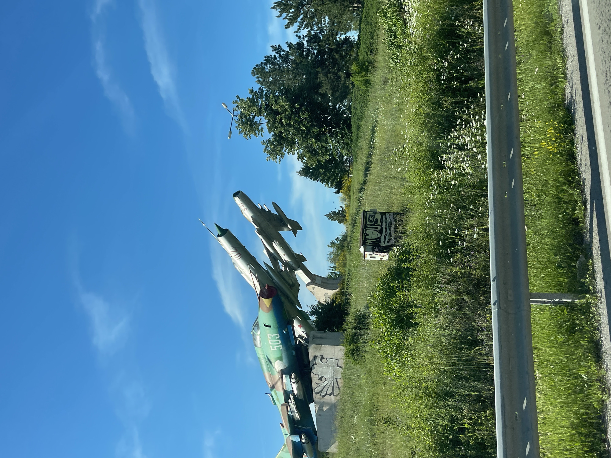 An outdoor, medium-shot photograph captures a monument to two Soviet-era fighter jets, a MiG-21 and a MiG-17, positioned on a grassy hill in Omurtag, Bulgaria. The jets are painted in a camouflage pattern of green, brown, and black, with the MiG-21 prominently displaying the number "503" in white. Both aircraft are mounted on concrete pedestals, with the MiG-21 elevated and angled towards the viewer's left. The surrounding environment is characterized by a vibrant green, grassy slope dotted with small white flowers. Tall green trees, including coniferous varieties, form a backdrop on the right side of the frame, with a clear blue sky featuring scattered white clouds above. In the foreground, a grey metal guardrail runs horizontally across the bottom of the image, separating the viewer from the grassy embankment and the monument. To the right and slightly in front of the monument, a small, weathered, rectangular structure with graffiti on its sides is visible. The scene appears to be bathed in daylight, suggesting a sunny day.