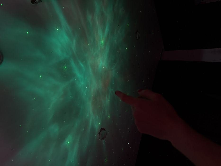A green nebula-like projection illuminates a wall, sprinkled with small, glowing points resembling stars. A hand extends from the right side of the frame, with the index finger pointing towards the center of the nebula. Circular fixtures are visible on the wall, suggesting light sources or other architectural elements. The right side of the image is predominantly dark, contrasting with the vibrant projection on the wall.