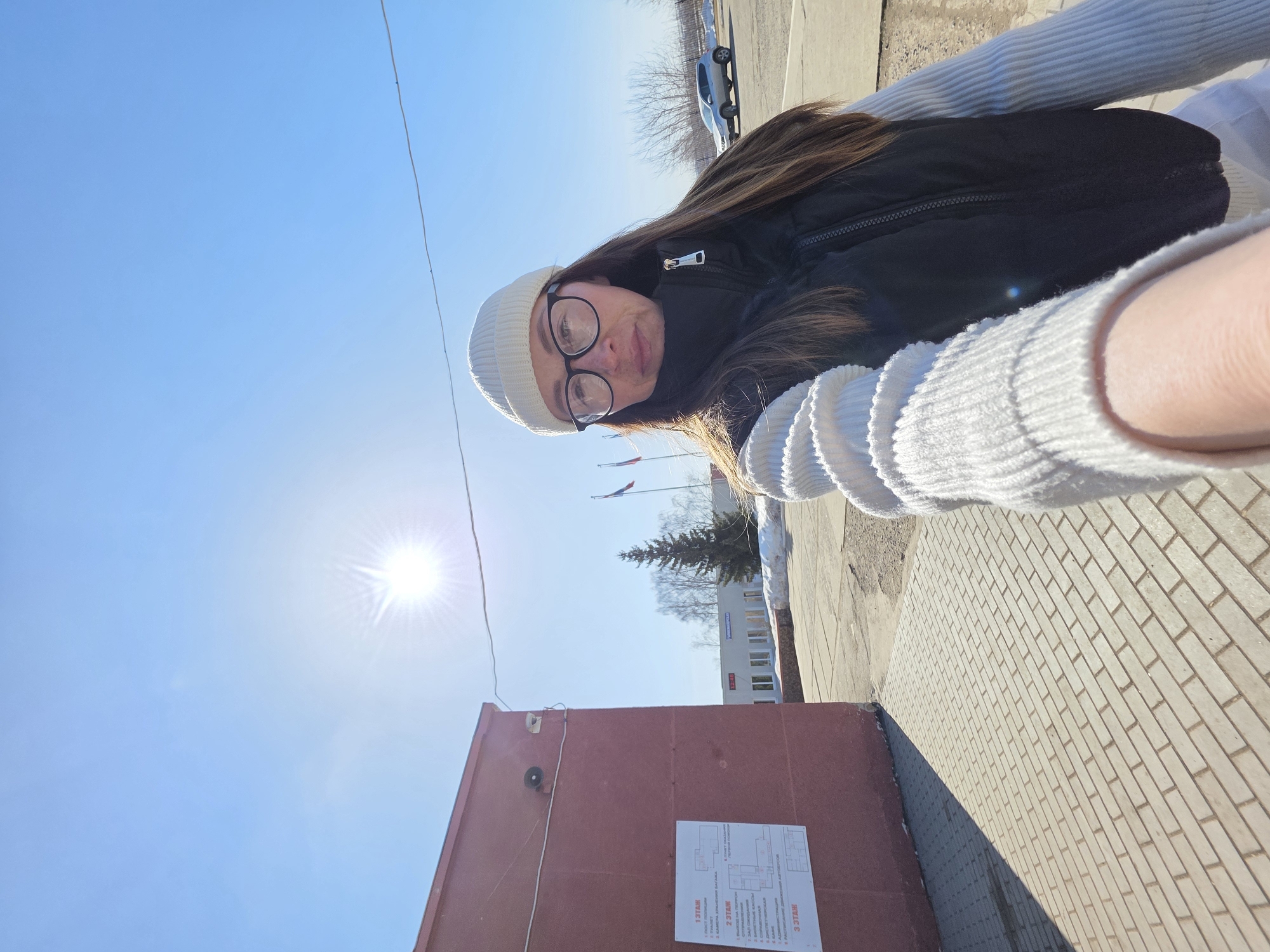 The image is a selfie taken by a woman wearing glasses and a white beanie, a black vest, and a white sweater. She is outdoors on a sunny day. The perspective is tilted, looking down on the scene.
In the background, there is a building with Russian flags. A brown wall is on the left side of the image with a posted diagram on it. There is a brick path visible.
The location is likely in the city of Chaikovsky, Russia, given the presence of Russian flags.