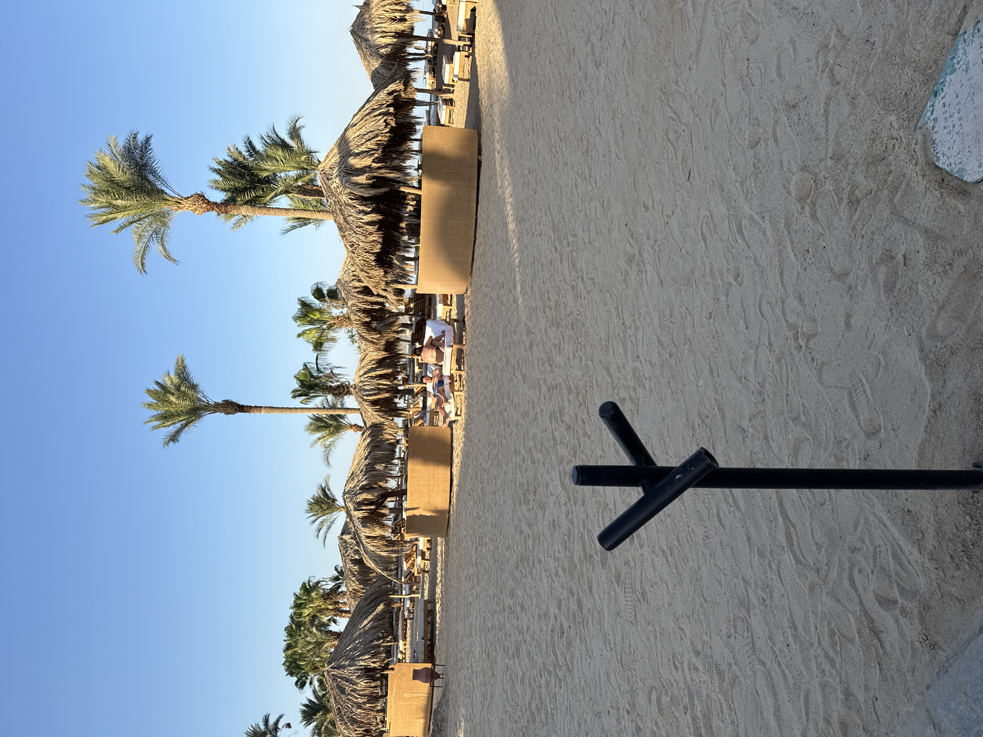 The image depicts a sunny beach scene in Hurghada, Egypt. The foreground is dominated by a sandy beach with visible footprints and a dark, Y-shaped metal post. In the mid-ground, there are several thatched-roof cabanas and lounge chairs, with some people relaxing. Palm trees are scattered throughout the scene, and the sky is clear and blue. The environment suggests a resort or hotel area on the Red Sea coast.