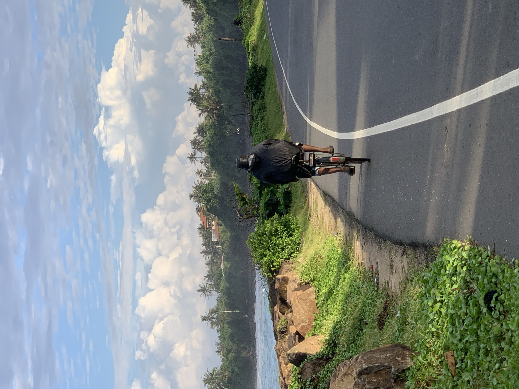 The image depicts a man cycling on a paved road in Kathaluwa, Sri Lanka. The man is seen from behind, wearing a dark hooded jacket and shorts, and a bucket hat. He is riding a bicycle with a rear rack. The road curves to the right, bordered by a grassy shoulder on the left. Beyond the shoulder, there are large rocks, lush green vegetation, and the ocean. The coastline is lined with palm trees and dense foliage, with some houses visible on a hill in the distance. The sky is filled with white, puffy clouds against a blue backdrop. The overall scene suggests an outdoor environment, likely a coastal area, with a focus on the man's solitary journey on the road.