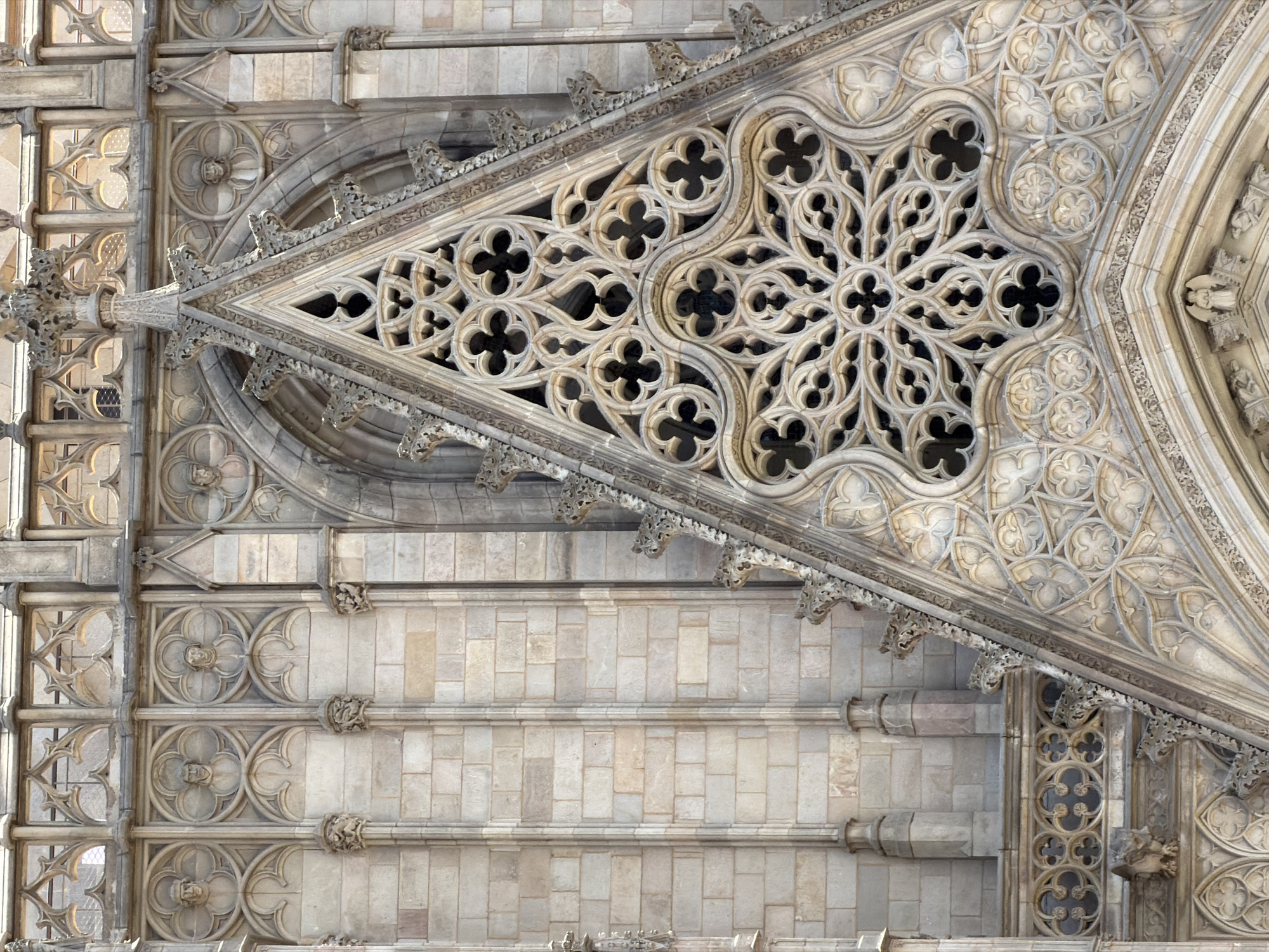 The image is a close-up, detailed view of the facade of the Barcelona Cathedral in Barcelona, Spain. The focus is on the intricate Gothic architectural elements of the building, specifically a large, ornate rose window and surrounding stonework. The rose window, centrally positioned, is a complex geometric pattern of carved stone with multiple trefoil and quatrefoil motifs, creating a starburst effect. Below the rose window, the facade features layered architectural details, including smaller decorative tracery, moldings, and what appear to be carved faces or figures within circular frames on the left side of the image. The stonework is weathered and shows the characteristic light beige and grey tones of aged sandstone or limestone. Vertical fluted columns and brickwork are visible to the left, providing a contrast in texture and pattern. The overall impression is one of meticulous craftsmanship and historical grandeur.