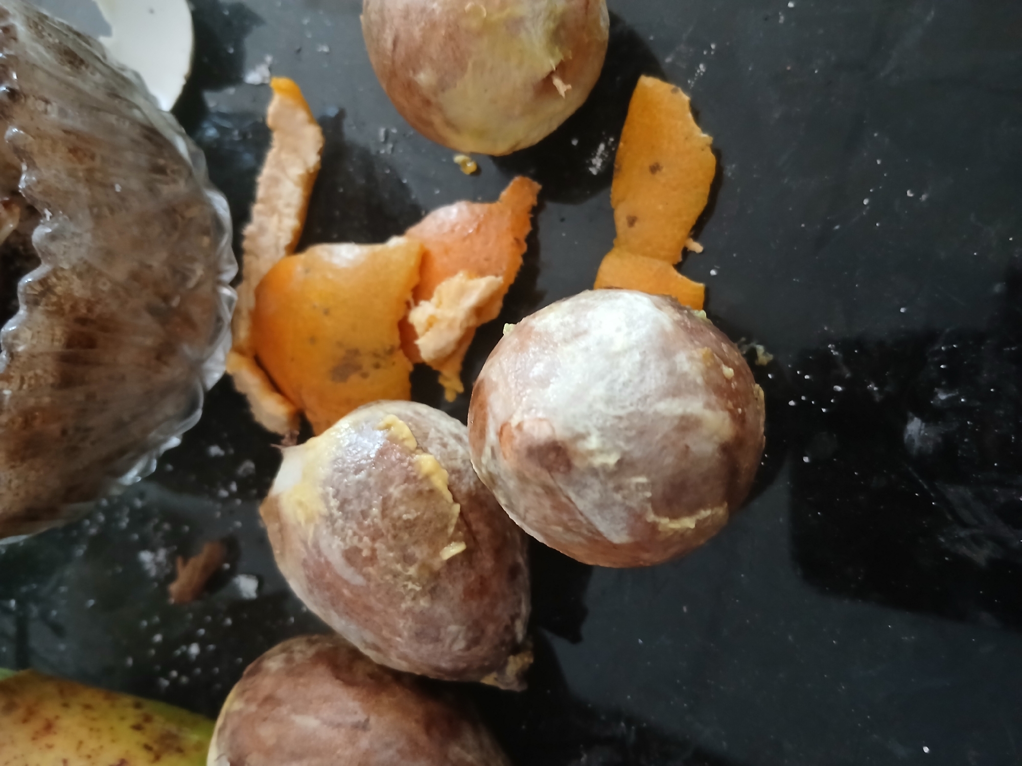 The image shows avocado pits scattered on a dark, reflective surface, possibly a countertop. Some of the pits have remnants of avocado flesh clinging to them, appearing creamy white and yellow. There are also pieces of orange peel interspersed among the pits, suggesting a meal or preparation that involved both fruits. In the background, a blurred, clear glass bowl is visible, containing what appears to be a mixture of dark grains or seeds. A portion of a yellow-green fruit, possibly a mango, is partially visible on the far left. The overall scene is domestic and appears to be indoors. The image does not contain any information about a specific location in Tenggarang, Indonesia, or any people.