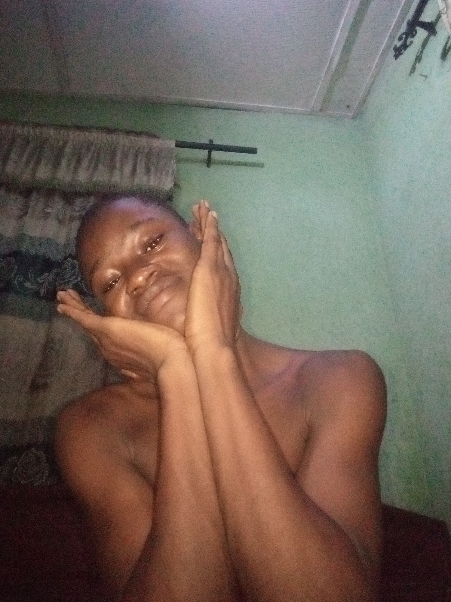 A selfie, taken indoors, shows a person with dark skin and short hair resting their head on their hands. The person is looking at the camera with a soft expression. Their arms are bent, with hands framing their face. The background is a plain green wall, with a section of patterned fabric visible behind the person's head and shoulders. A dark ceiling is visible at the top of the image. The lighting is dim, creating shadows on the person's face and body. The image appears to have been taken in a residential setting. This image was taken in Shangisha, Nigeria.
