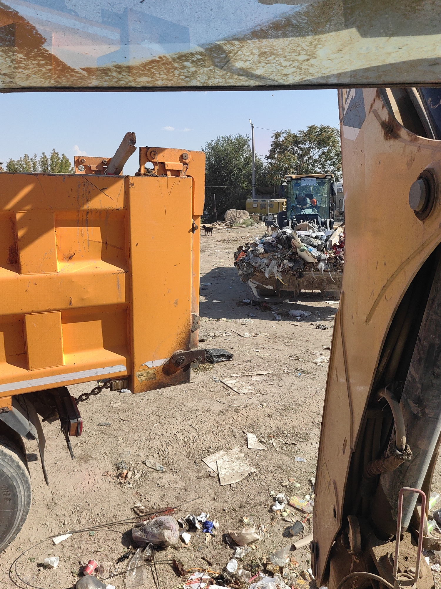 The image depicts a landfill site, likely in or near the city of Shymkent, Kazakhstan. In the foreground, the orange body of a dump truck is visible, occupying the left half of the frame. Its side is marked with the typical industrial paint and signs of wear. To the right, a large, orange construction machine, possibly a loader or excavator, is positioned close to the dump truck, with its arm and bucket visible. The bucket of this machine is filled with a large quantity of mixed waste, including plastic bags, paper, and other refuse.
In the middle ground, a yellow backhoe or similar heavy machinery is operating, with a person visible inside the cab. This machine is also near piles of garbage. The ground is uneven and covered with dirt, debris, and scattered trash, characteristic of a waste disposal area. A small black dog is visible in the middle distance, standing on the dirt.
The background shows a line of trees and vegetation, with a clear blue sky overhead. A tall, thin pole, possibly a lamppost, is also present. The overall environment suggests an active, if somewhat unkempt, landfill operation. The presence of heavy machinery and a dump truck indicates ongoing waste management activities. The scene is illuminated by natural daylight.
