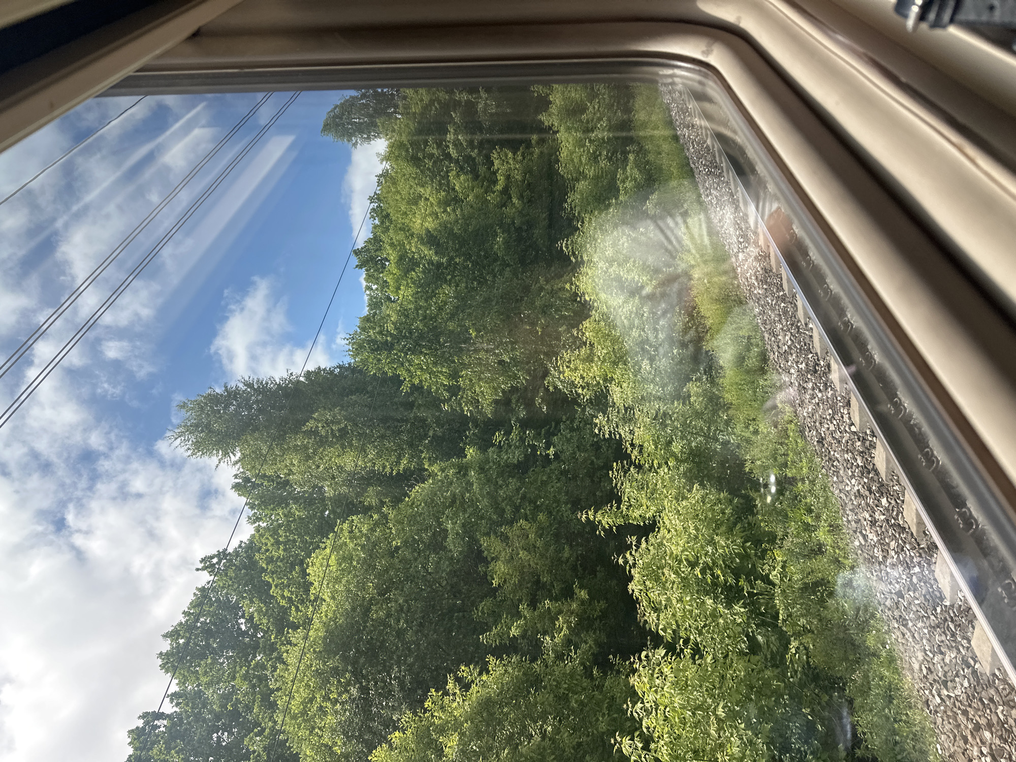 The image displays a vibrant view from inside a vehicle window, likely a train, showcasing a lush natural landscape under a bright sky. The light beige window frame is visible along the top and right edges, with reflections on the glass showing thin horizontal lines, possibly overhead power wires, and parts of the sky and clouds. A distinct sun flare is also present on the right side of the window.
Outside, the upper portion of the scene features a clear blue sky dotted with scattered white clouds. Below the sky, dense green trees with full foliage dominate the middle and left sections of the frame, suggesting a thriving forest or wooded area characteristic of spring or summer. Towards the lower right, parallel to the window, train tracks are visible, marked by grey gravel ballast and a concrete sleeper. A strip of vivid green bushes and undergrowth runs alongside the tracks. Several thin, dark lines, resembling power or catenary wires, stretch horizontally across the sky and through the treetops.
The scene conveys a sunny daytime journey through a verdant, natural environment, consistent with a rural or semi-rural setting like Куркачи, Russia. No people or visible text are present.