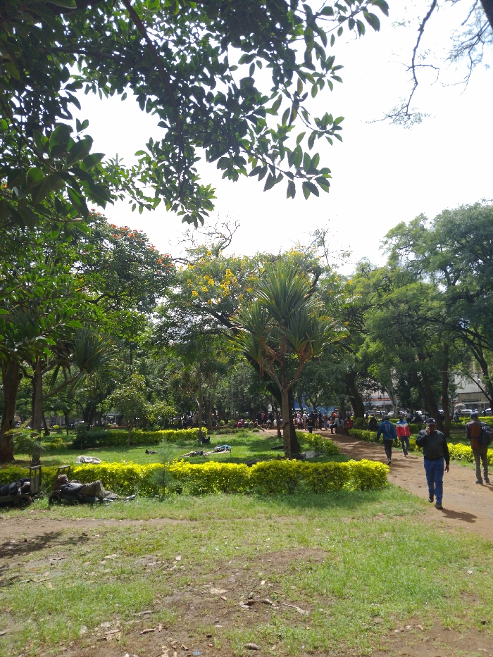 The image shows a park located at coordinates -1.453, 37.007 in the city of Athi River, Kenya. The park is characterized by green grass and various species of trees, including palm trees. There are several people walking on a path, and several more people are sitting and lying on the grass. Some of the people are wearing casual clothing, and some are wearing uniforms. The scene is peaceful and quiet. The weather is sunny and bright.