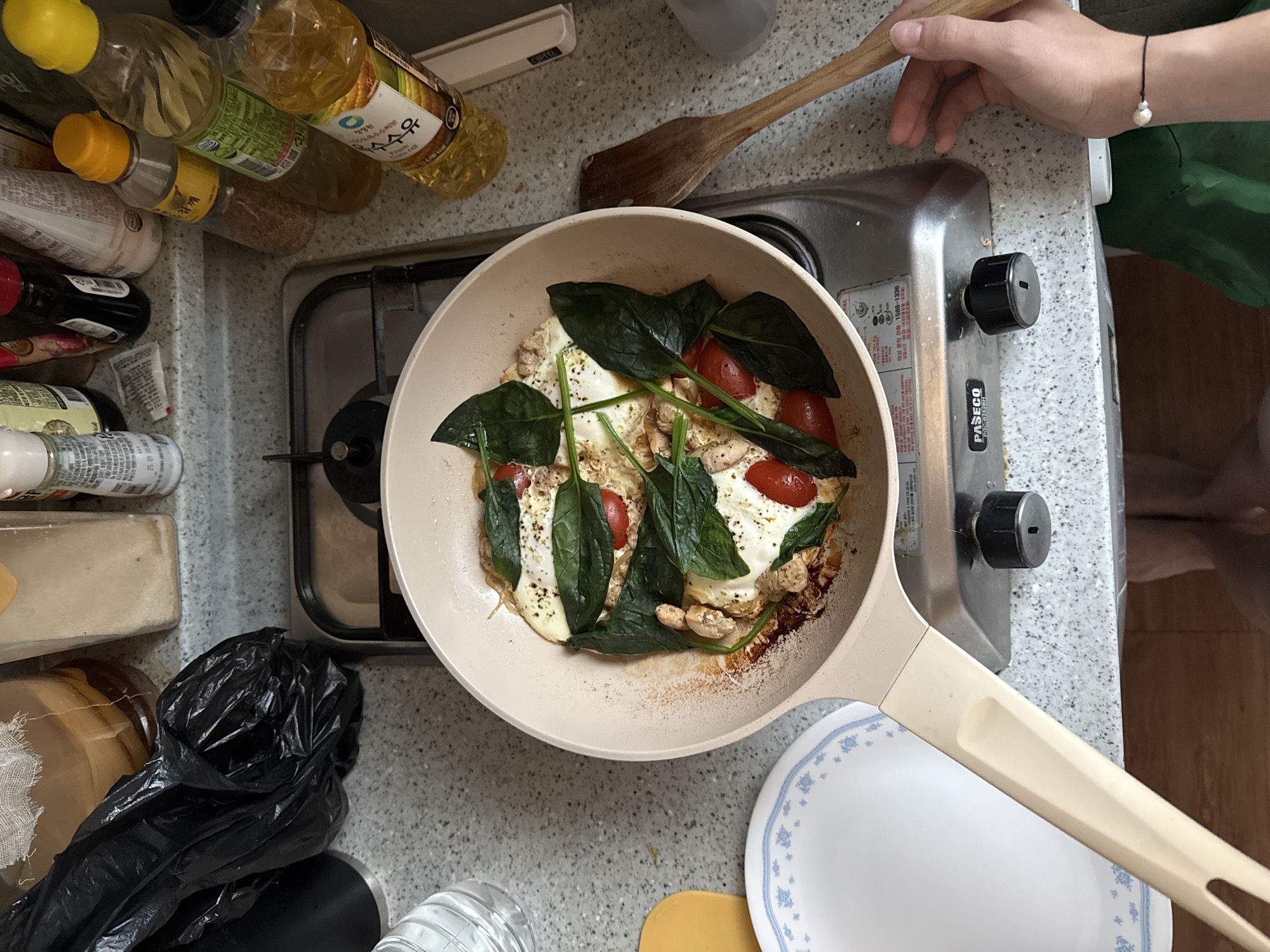 A top-down, close-up shot shows a person cooking in a kitchen, possibly in Gwangju, South Korea. The main focus is a light-colored pan on a gas stove, containing a cooked dish. The dish appears to be eggs, spinach, and cherry tomatoes. The pan is slightly browned on the edges, indicating it has been used for cooking. The gas stove is silver and has black knobs.
To the left of the stove, a white plate with a blue floral pattern is visible. Further left, a black trash bag sits on a speckled countertop. Behind the stove, various bottles of cooking oil and seasonings are arranged on a shelf, including a prominent bottle of corn oil with Korean writing. A wooden spatula is held by a person's hands on the right side of the frame, positioned near the pan. The person is wearing a bracelet with a single pearl. The background suggests a typical home kitchen environment with tiled countertops. The lighting is consistent with indoor lighting.