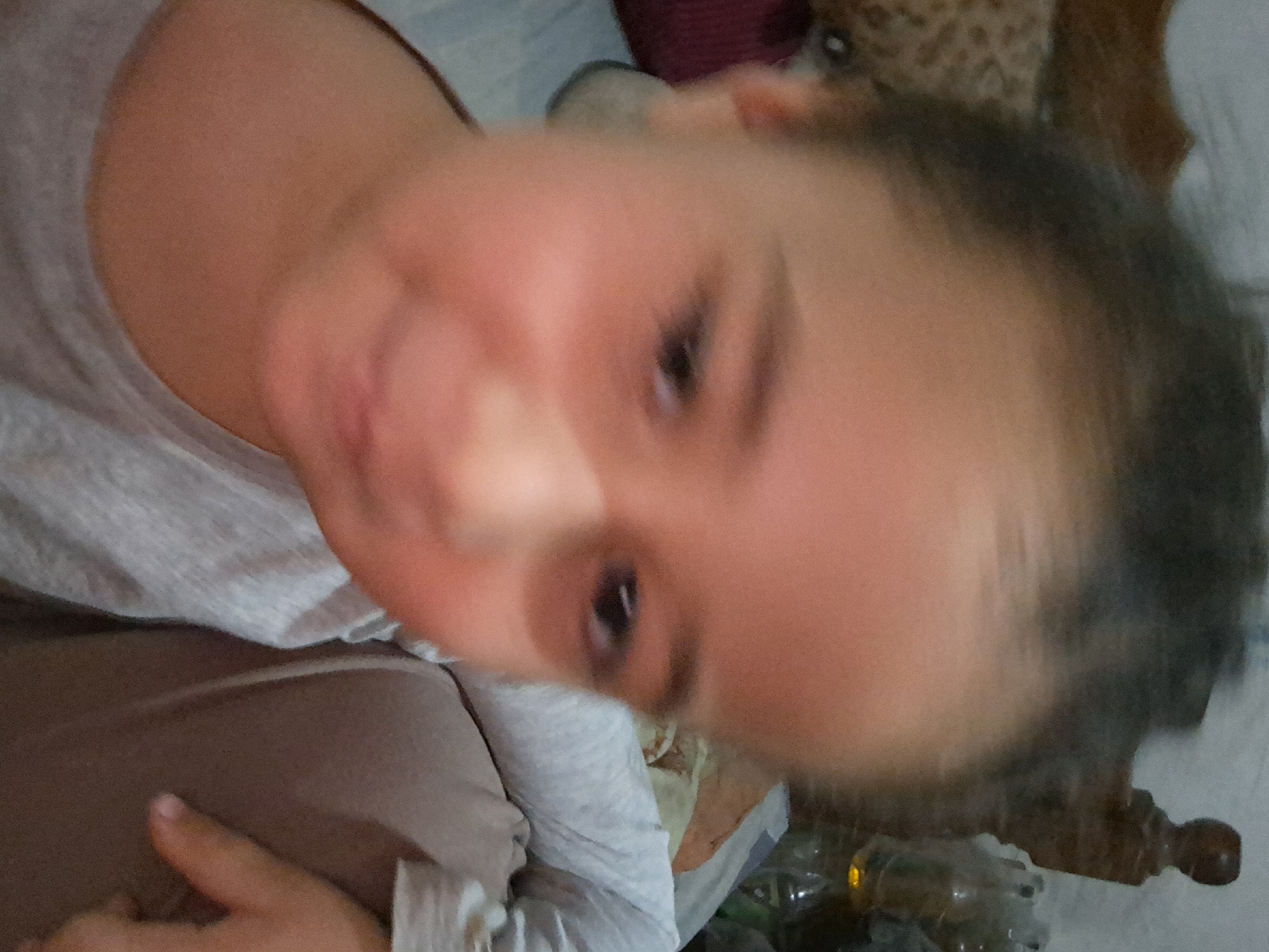 A young child is looking upwards towards the camera. They have dark hair, fair skin, and are wearing a grey shirt. The background includes a bed with white sheets, and some blurry objects that are difficult to discern. The location of the image, Juan Griego, Venezuela, cannot be confirmed from the visual data.