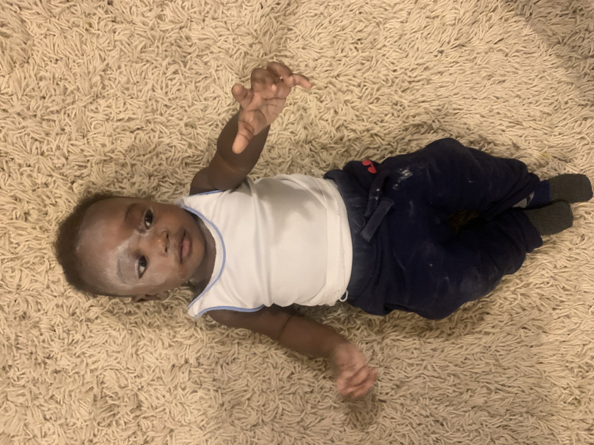 A baby lies on a cream-colored shag rug. The baby is wearing a white tank top with blue trim, dark blue pants, and gray socks. The baby's face has white powder or residue on it. The baby is lying on its back, with one arm raised. The location is not discernible as being Barakin Akawo, Nigeria.