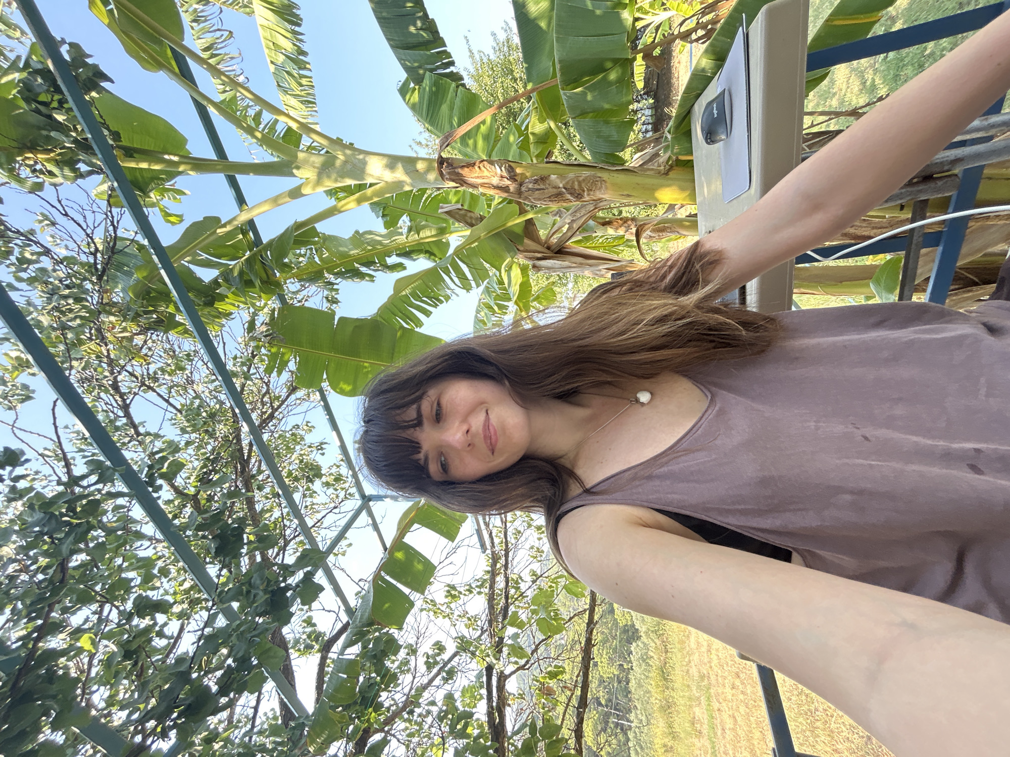 A woman with long brown hair and bangs is looking down and to the right, with a slight smile. She is wearing a light purple tank top over a black bra and a silver necklace with a white pendant. Her left arm is resting on a table. On the table, there is a laptop and a computer mouse on a mousepad. The woman is outdoors, with a large banana tree to her right and foliage to her left and above her. The background shows a field of dry grass and trees under a clear blue sky. The scene appears to be in a tropical or subtropical outdoor setting, possibly a balcony or patio. The image doesn't provide specific details to pinpoint the location within Ortaca, Turkey.