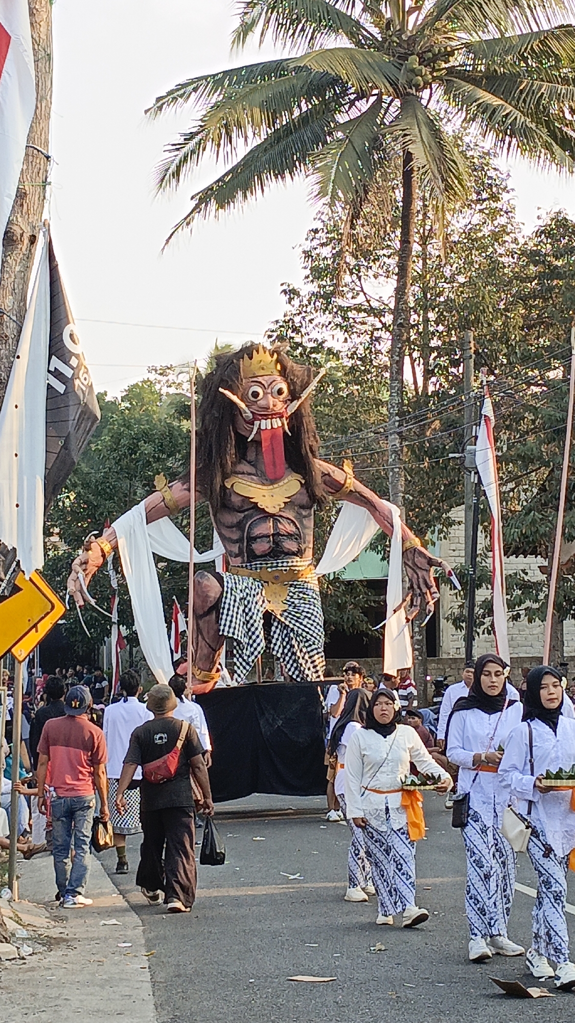 In Krajan Lor, Indonesia, a large, grotesque effigy of a demon-like figure with bulging eyes, a protruding tongue, and sharp claws is the central focus of a procession. The effigy, adorned with a crown and draped with white fabric, stands on a raised platform surrounded by black cloth. A crowd of people, some in traditional white attire and others in casual clothing, line the paved street. Several individuals are carrying offerings in woven baskets. Flags, including one with red and white stripes, are displayed on poles. Palm trees and other vegetation are visible in the background, beneath a bright sky. The scene appears to be a cultural or religious event, possibly a parade or festival.