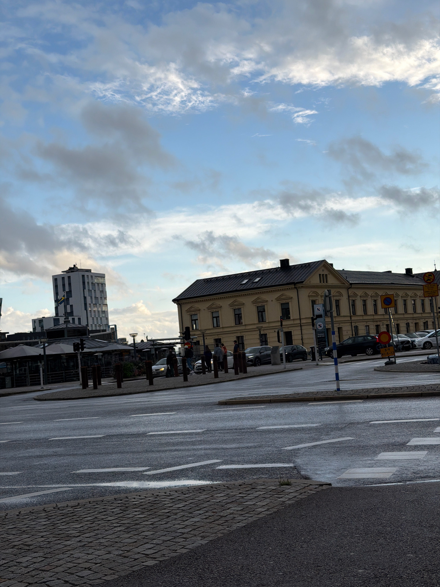 The image depicts a street scene in Helsingborg, Sweden, on a cloudy day. In the foreground, a wet asphalt road with white lane markings and a cobblestone sidewalk is visible. A crosswalk is present. In the mid-ground, there are several vehicles, including cars and what appears to be a white van or bus, on the road. Pedestrians are seen walking on the sidewalk. Behind the road, there are buildings, including a modern, tall, multi-story building with a flag on it, and a more traditional, long, light-yellow building with a dark roof. Several streetlights and traffic signals are also present. The sky is filled with a mix of blue sky and gray clouds. The overall impression is that of an urban environment.