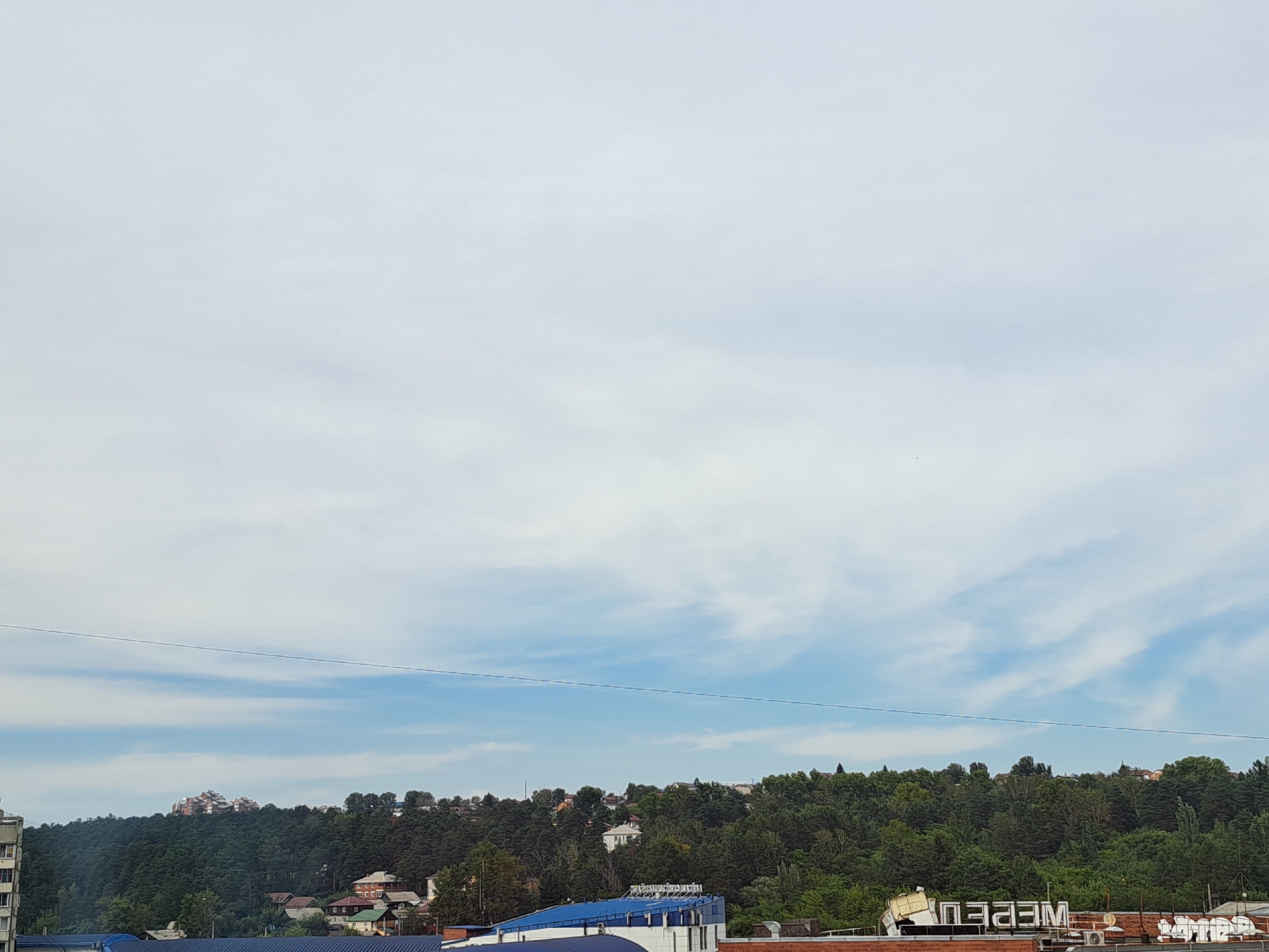 The image shows a slightly overcast sky with light blue and white clouds. In the middle ground, a densely forested hill rises, dotted with houses and buildings of varying architectural styles. A prominent cluster of multi-story buildings with red roofs sits atop the hill, suggesting a residential development. Below the hill, a scattering of smaller houses with pitched roofs are visible amongst the trees. In the foreground, the roofs of several industrial or commercial buildings are apparent. One building has a bright blue roof and a white structure with what appears to be ventilation units on top. Another building has a large, partially obscured white structure with dark lettering that is backward, possibly indicating a reflection or a sign for an establishment. The scene captures a view of a hillside neighborhood with commercial structures in the foreground, typical of a city edge. It is not possible to ascertain the specific location within Irkutsk, Russia from the visual information provided. There are no people or discernable actions depicted in the image.