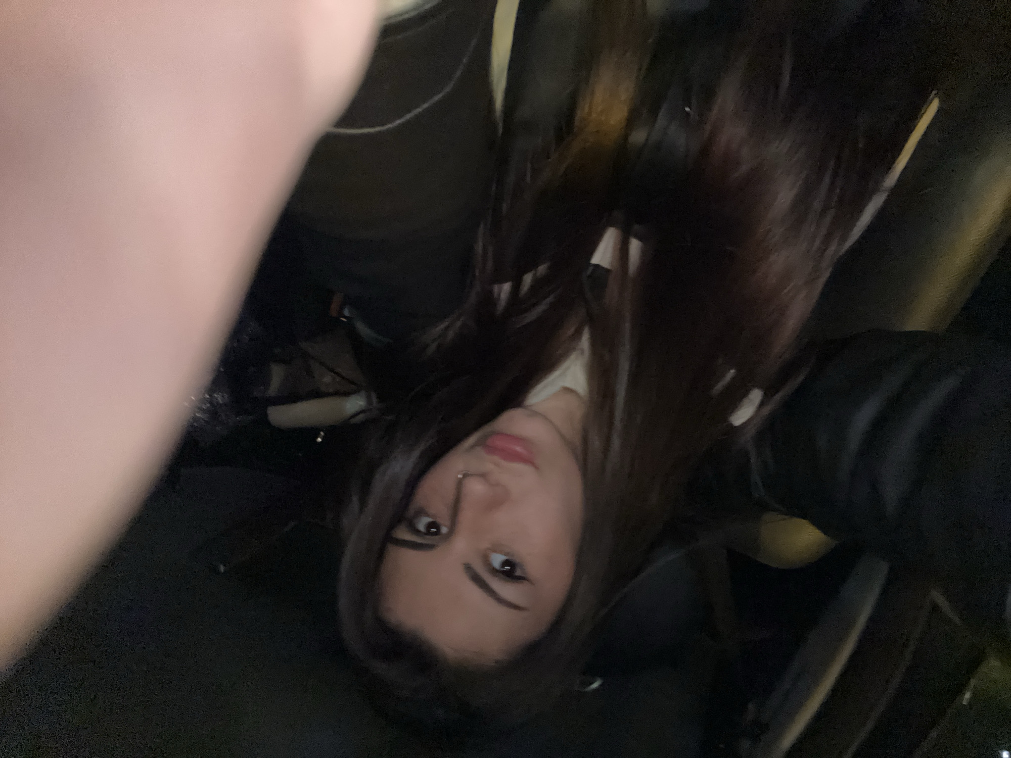 A person is in a vehicle, possibly a car, as evidenced by the car seat and interior visible in the background. The subject has long, dark hair, dark eyes, and a nose piercing. They appear to be wearing a white top and a dark-colored jacket. The photo is taken from an unusual angle, potentially a selfie. The image does not provide any information to ascertain the location.