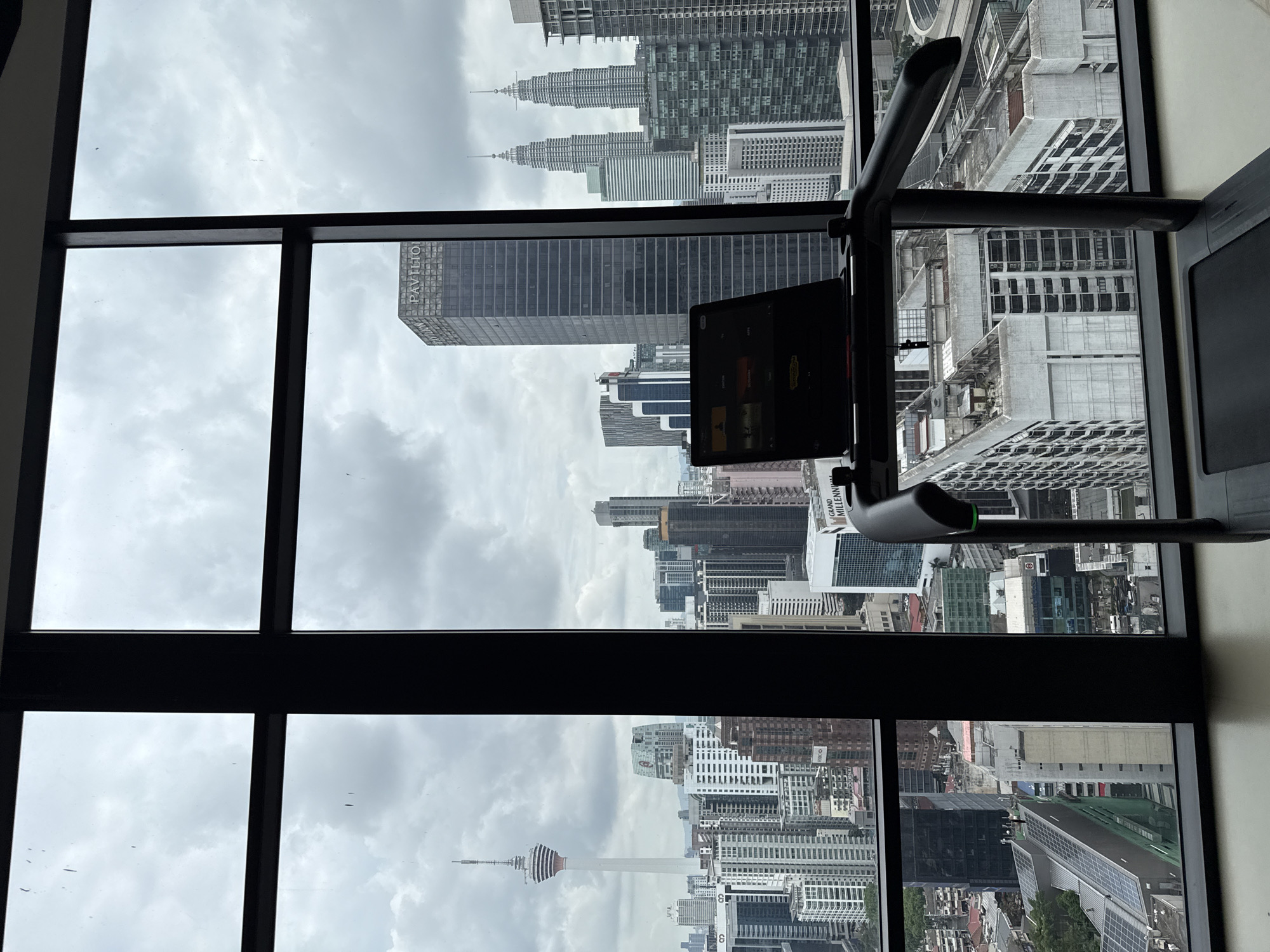 The image shows a view of Kuala Lumpur, Malaysia through a window, possibly from a high-rise building. A piece of exercise equipment is visible on the right. Landmarks are seen in the distance, including the Petronas Twin Towers and the Kuala Lumpur Tower (KL Tower). The weather appears overcast, and the city consists of high-density urban development with numerous skyscrapers.