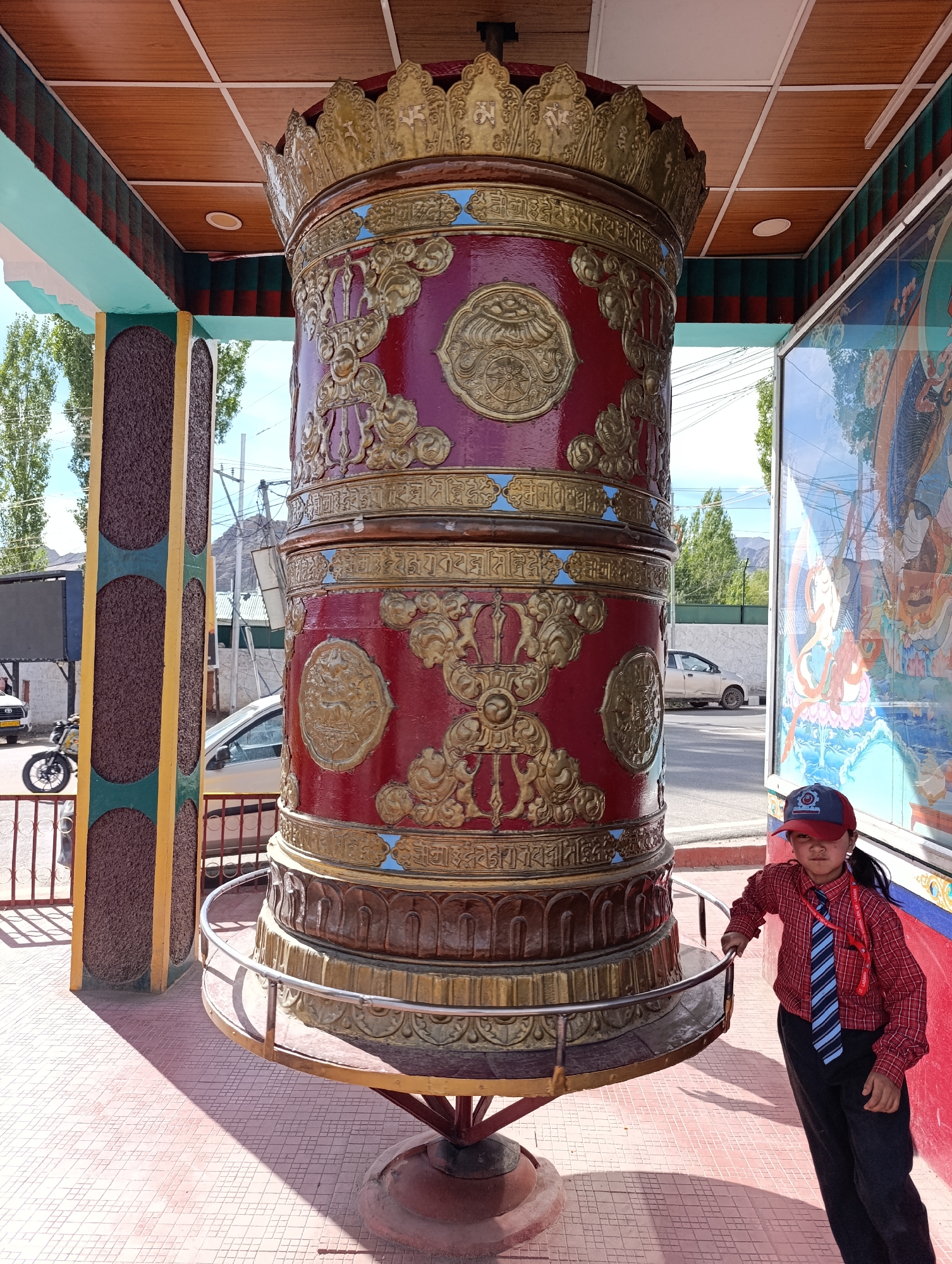 In the city of Leh, India, a young girl in a red plaid shirt, blue and white striped tie, and a red baseball cap stands next to a large, ornate red and gold prayer wheel. The prayer wheel, adorned with intricate gold carvings and Tibetan script, is mounted on a stand and surrounded by a metal railing. The girl's left hand rests on the railing. Behind her, a colorful mural depicting figures is visible on a wall. The scene is outdoors, with a paved ground and trees in the background. The sky is visible, indicating a daytime setting.