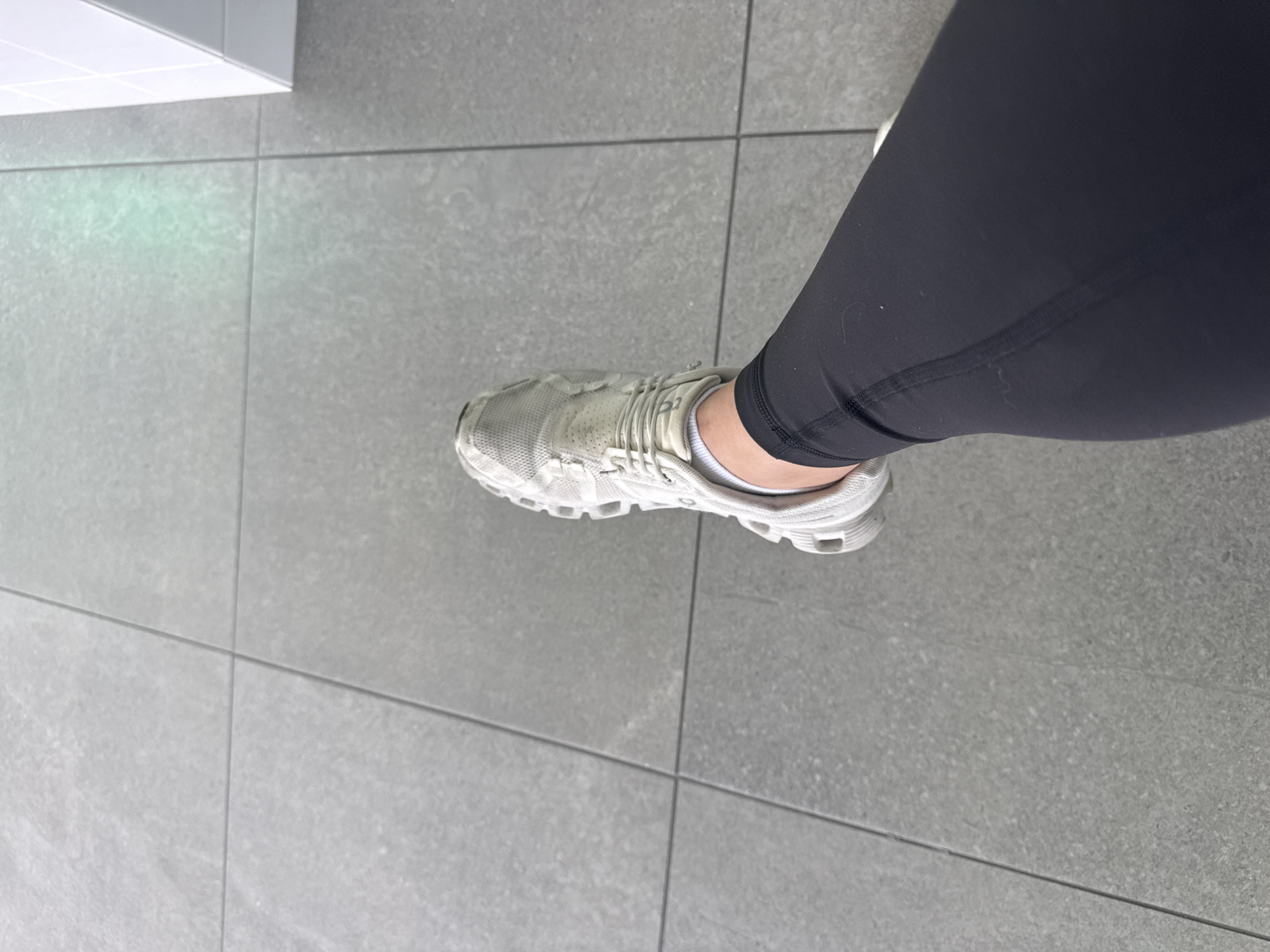 The image presents a close-up, high-angle view of a person's lower right leg and foot, positioned on a grey tiled floor. The person is wearing dark, likely black, athletic leggings that cover the leg down to the ankle, with white ankle socks visible between the leggings and the shoe. A light-colored (white or very light grey) athletic shoe, featuring a mesh-like upper texture and a distinct, chunky sole design, is on the foot. The floor is composed of large, rectangular grey tiles with clearly defined grout lines, suggesting an indoor setting like a gym, hallway, or modern commercial building. In the upper left portion of the frame, a white structural element, possibly a baseboard or part of a wall/cabinet, is visible, along with a faint, light green reflection on the floor, which could be from an overhead light fixture or screen. No specific activity is depicted other than standing, and there is no visible text. The image provides a detail-oriented view of the attire and immediate surroundings in what is contextually San Bernardino Tlaxcalancingo, Mexico.