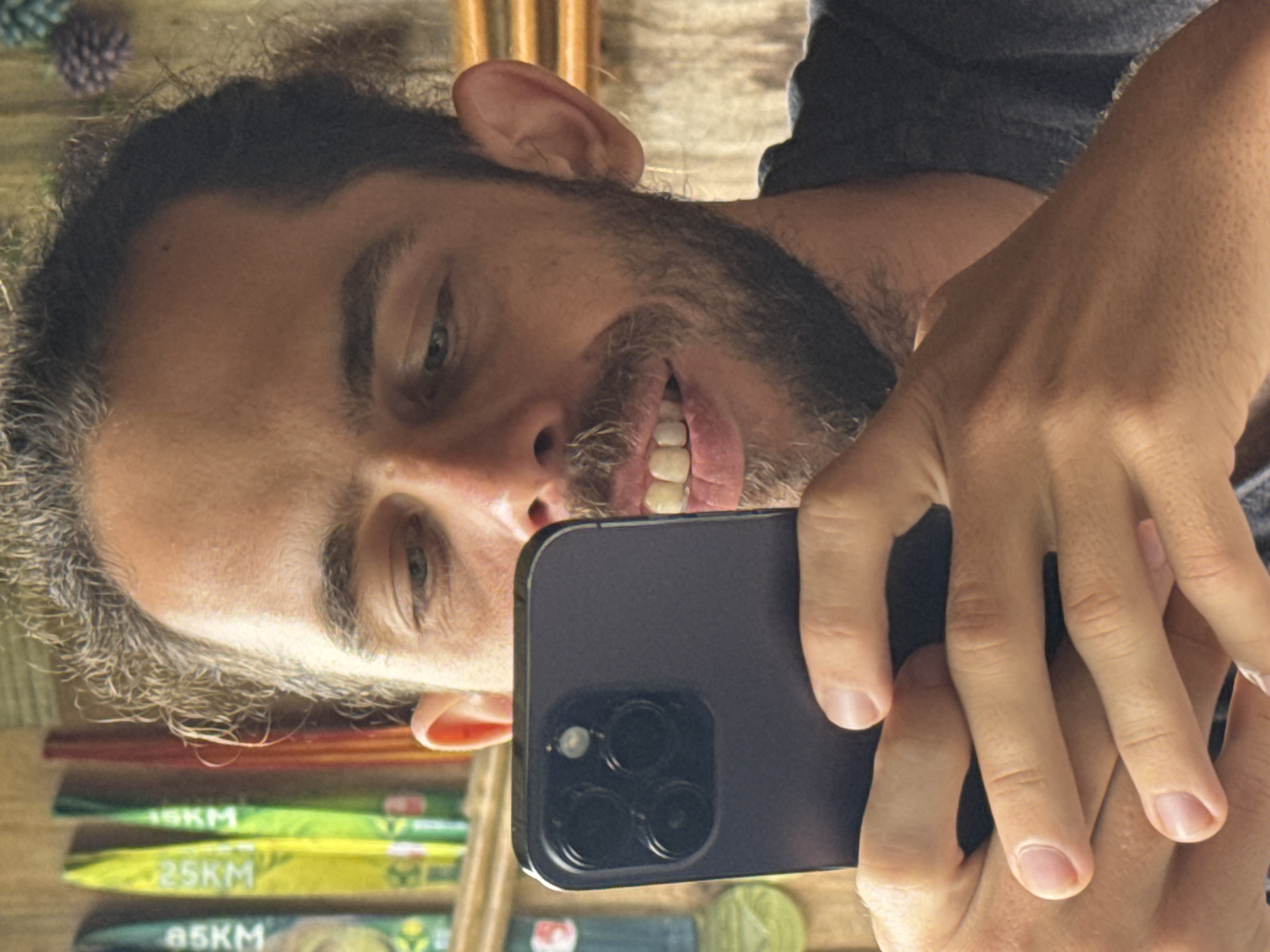 A man with graying dark hair and a beard is taking a selfie with a dark-colored smartphone. The phone has a triple-lens camera system. The man is smiling, showing his teeth. In the background, there are what appears to be nutrition bars or energy gels labeled with distances "15KM", "25KM," and "85KM." The location is presumably Ngã Ba Kim, Vietnam. The photo is taken in what seems to be an indoor setting.