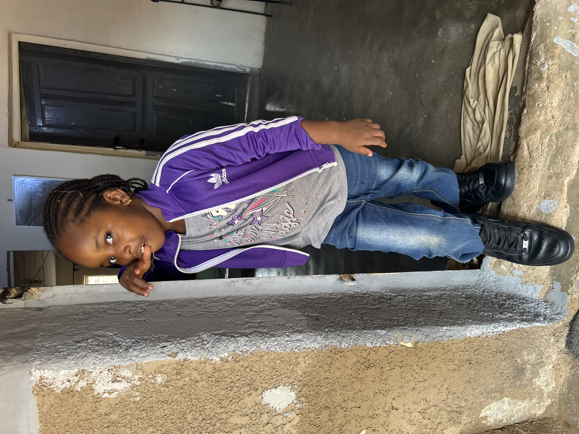 A young child is pictured standing on a gray and tan wall in what appears to be a doorway or opening in Jos, Nigeria. The child, wearing a purple Adidas jacket, gray shirt, and blue jeans, is looking at the camera and has a hand near their mouth. The child also wears black sneakers. The background includes a dark door, a concrete floor, and a beige cloth. The walls appear to be textured.